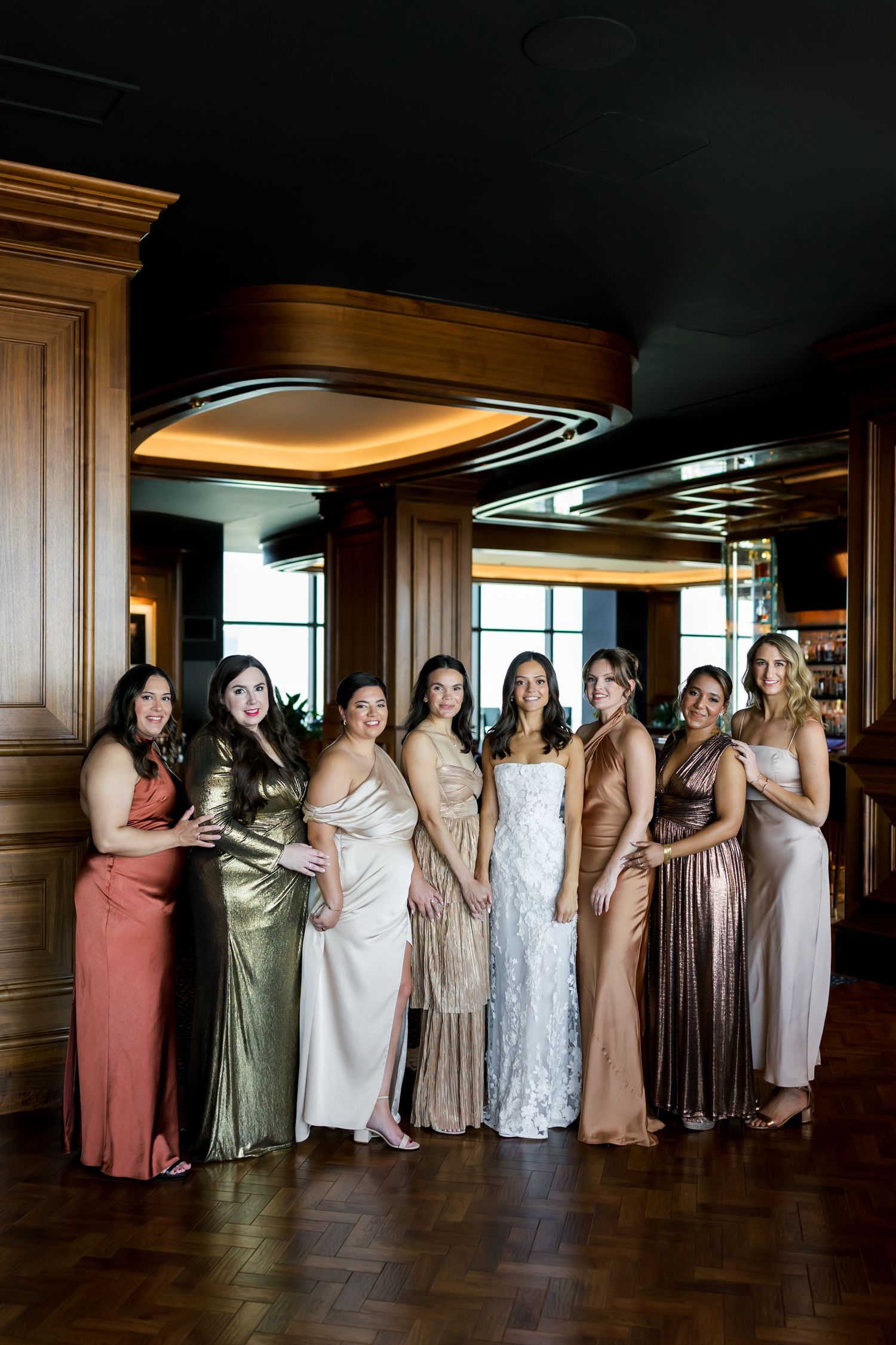 A bride in white lace gown poses with eight bridesmaids in metallic and neutral gowns inside an elegant wood-paneled venue.
