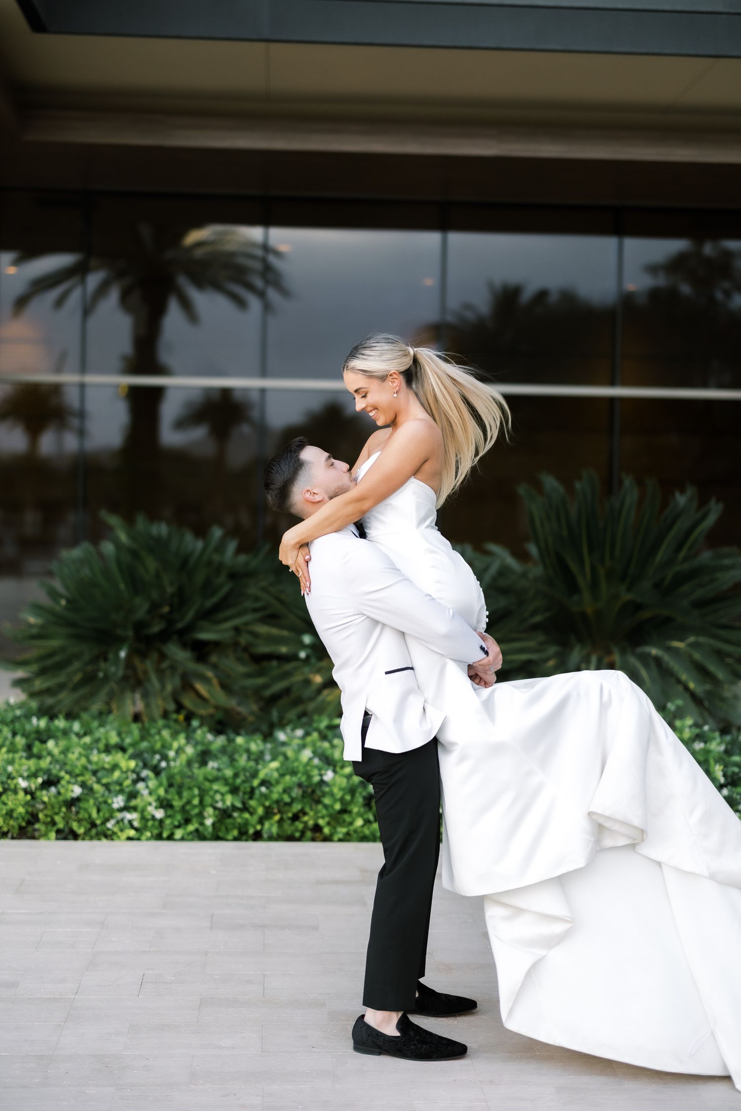 Groom in white tuxedo lifts smiling bride in strapless gown outside a glass building with palms.