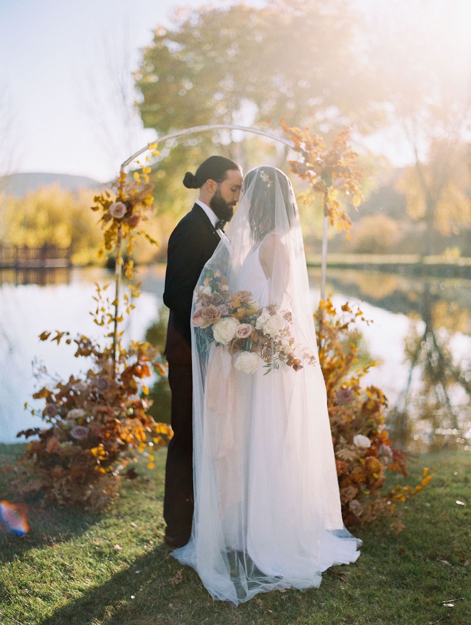 Bride and groom share a romantic moment at fall outdoor wedding ceremony by a lake with autumn floral arch backdrop.