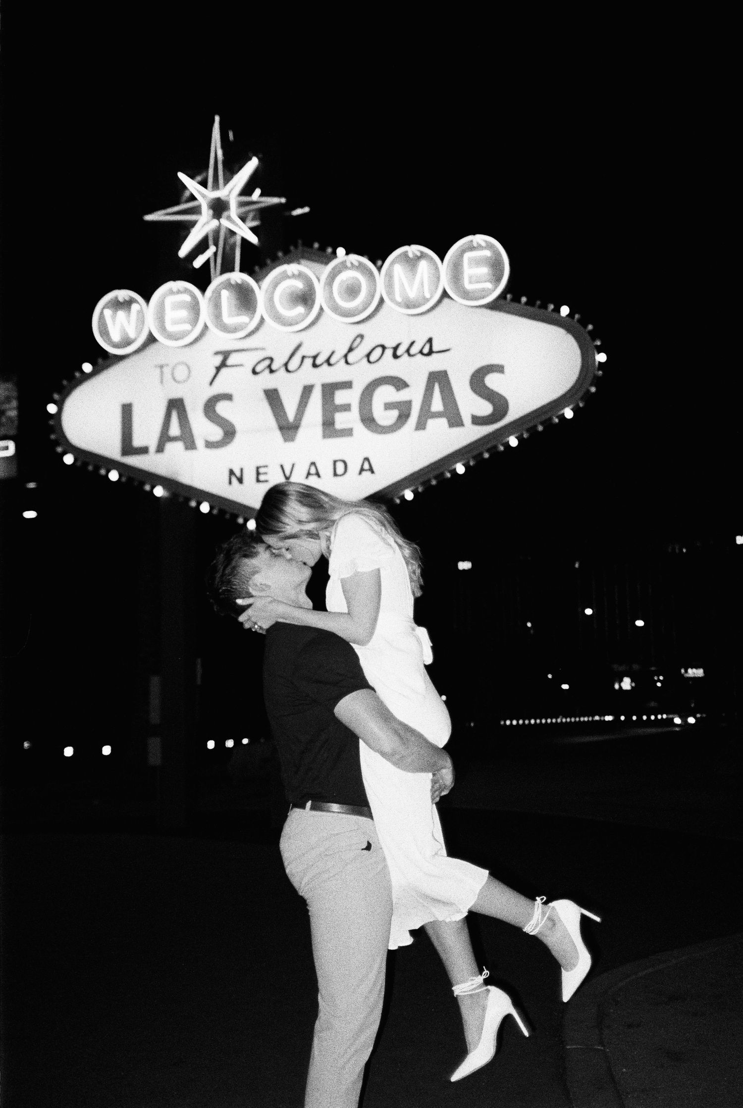 Couple kissing in front of the iconic Welcome to Fabulous Las Vegas Nevada sign at night, black and white photo.