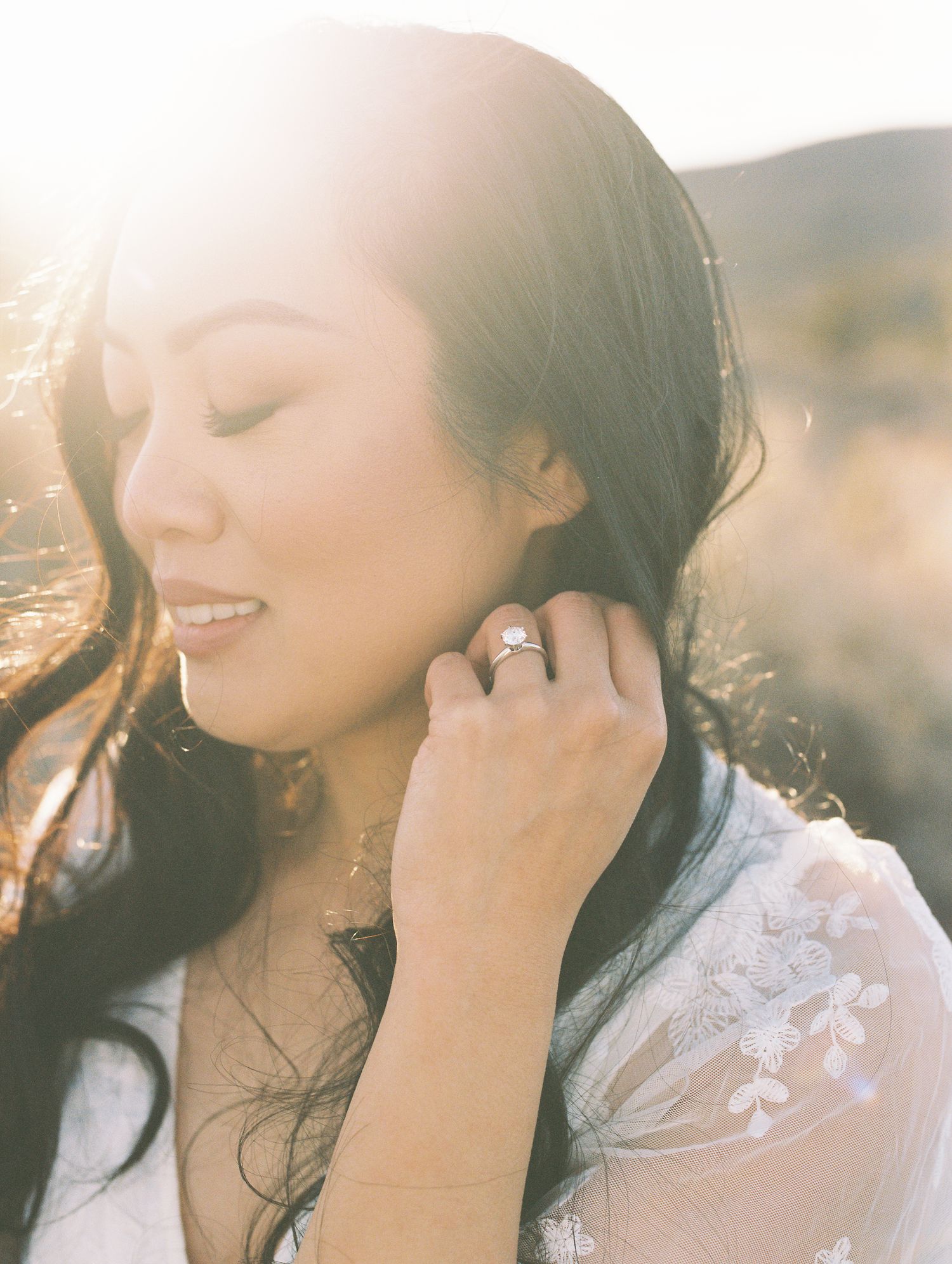 A bride with dark hair holds her ear, showcasing a stunning engagement ring, bathed in golden sunlight outdoors.