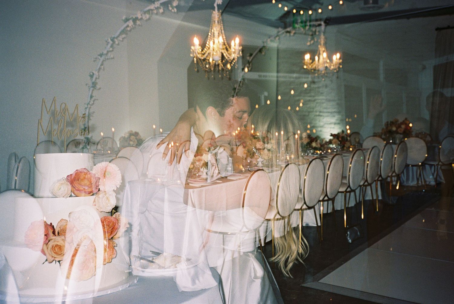 Elegant wedding reception with a floral white cake, long dining table, gold chairs, and warm chandelier lighting.
