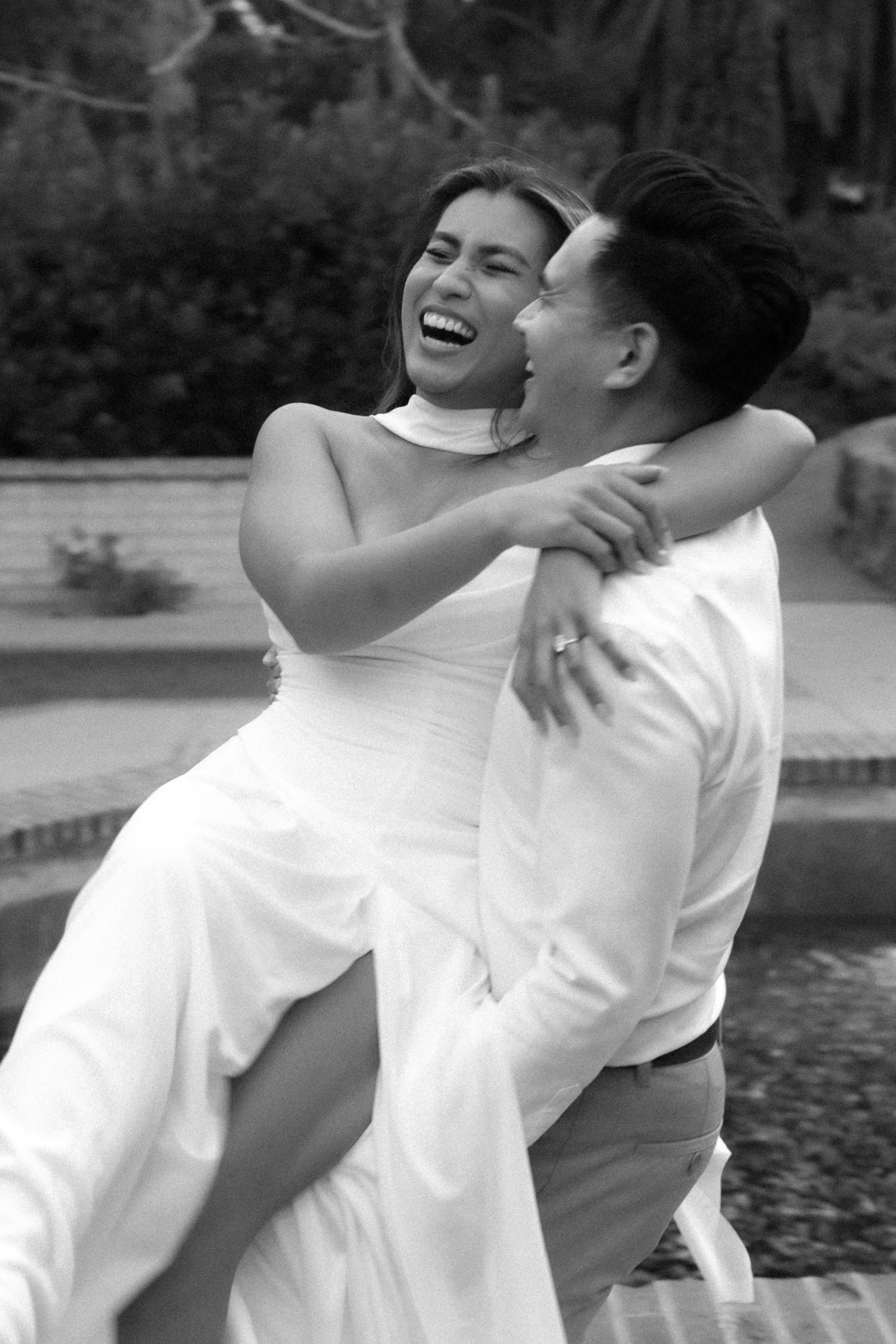 Black and white photo of a joyful bride being carried by groom, both laughing, in a garden setting.
