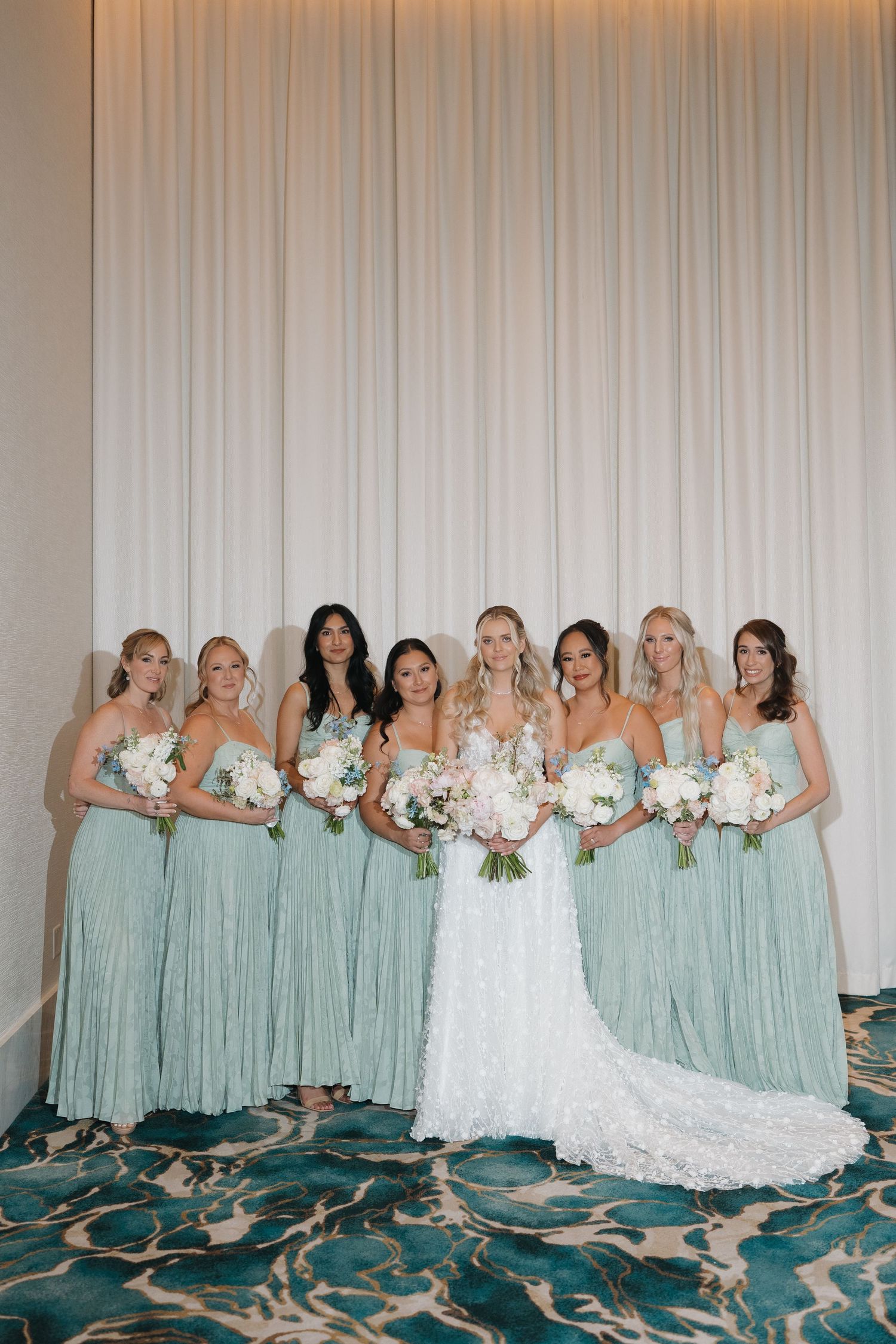 Bride with seven bridesmaids in sage green gowns holding white bouquets at wedding venue.