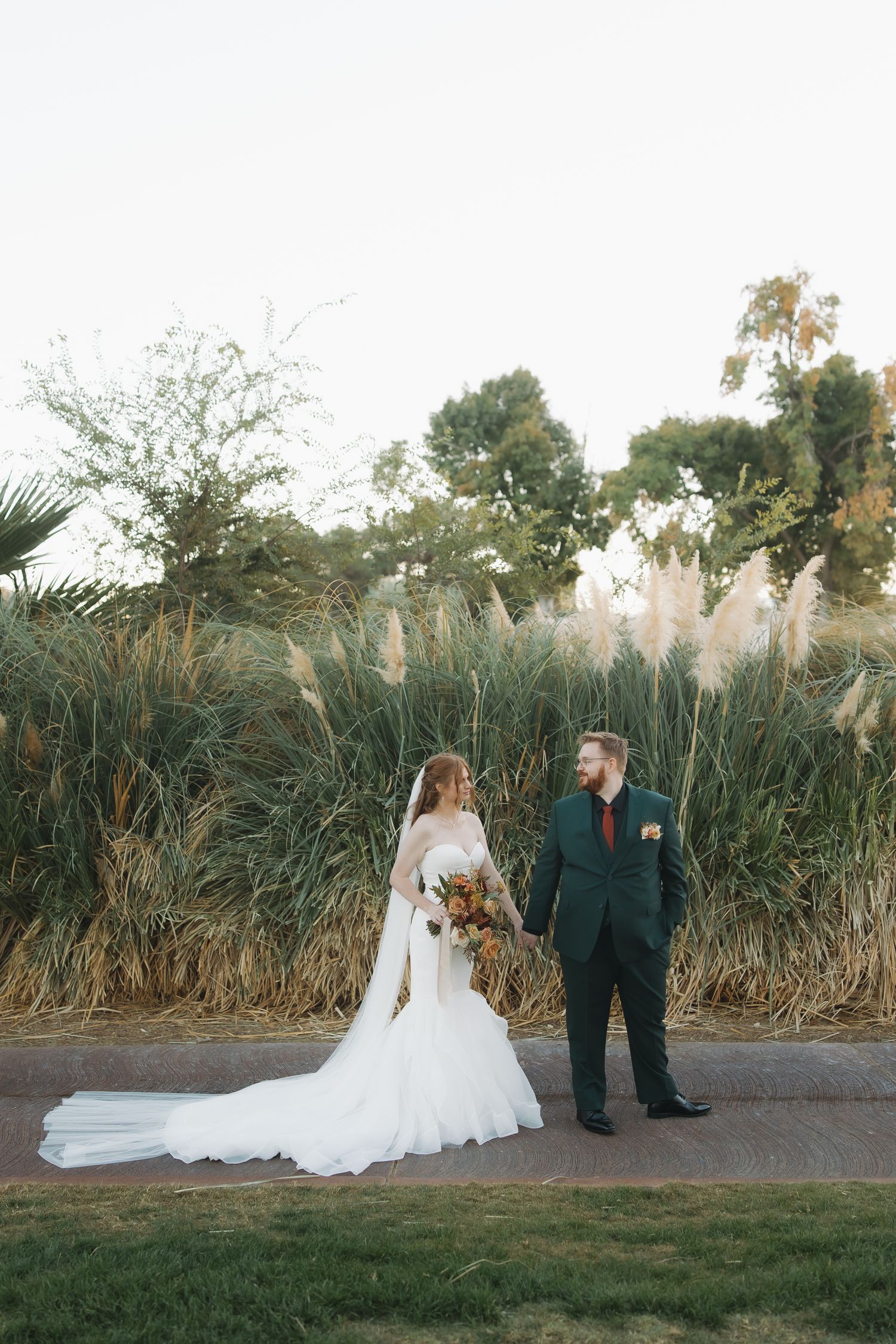 Bride in mermaid gown and groom in dark green suit hold hands before tall ornamental grass backdrop.