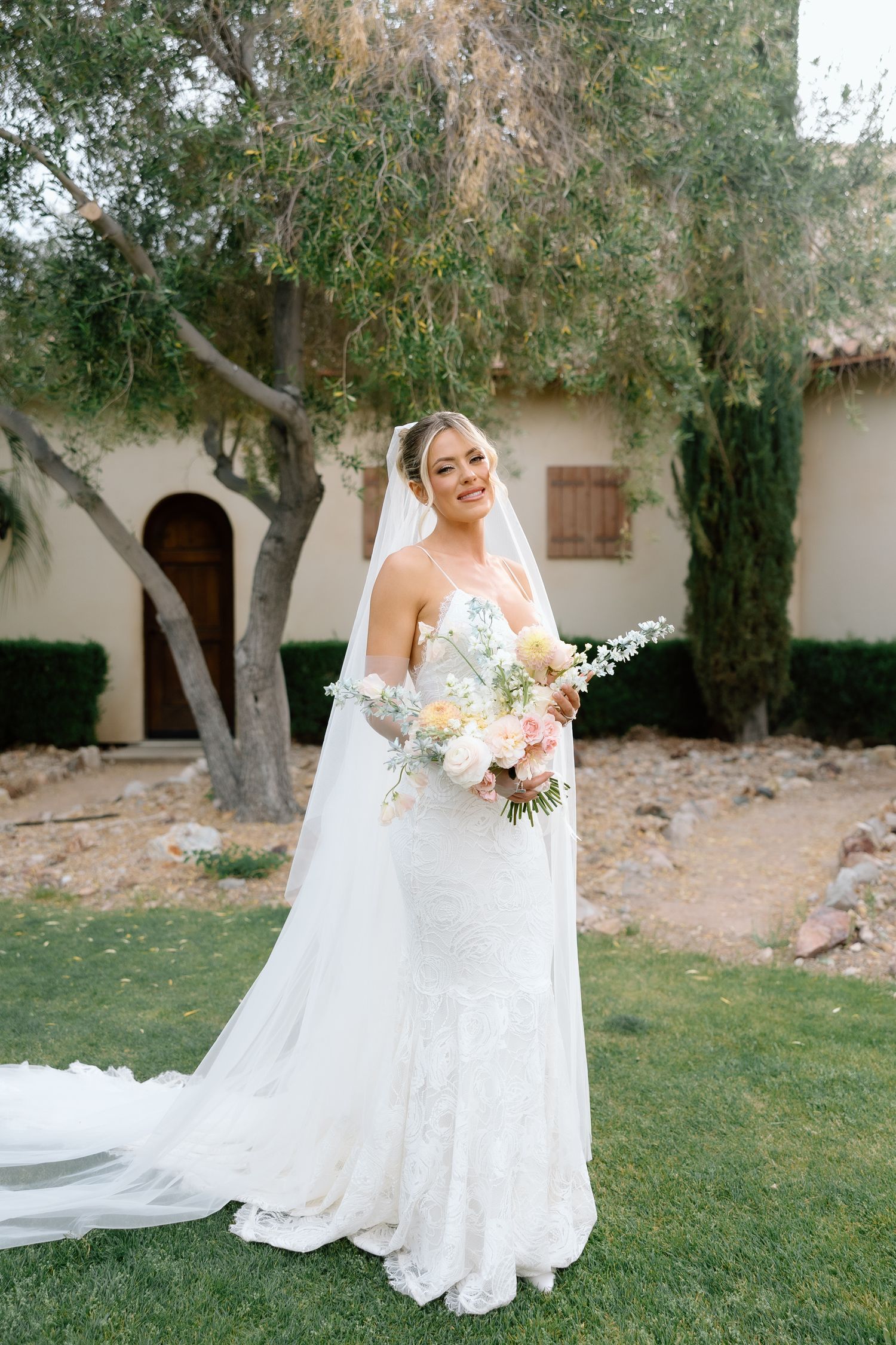 Bride in lace mermaid gown and veil holds pastel bouquet, posing outdoors near trees and Spanish-style building.