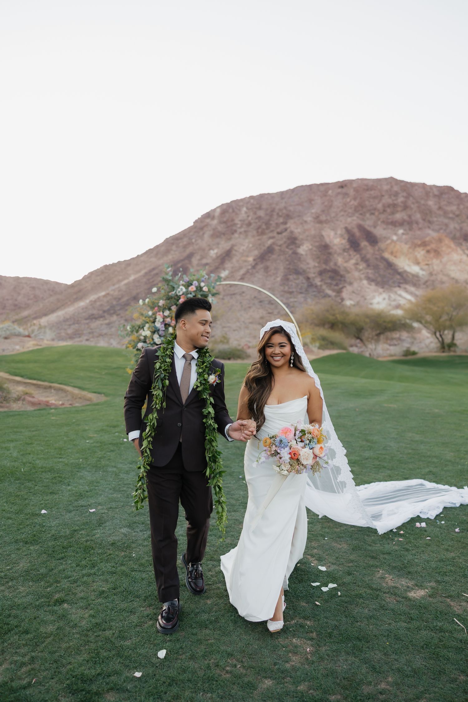 Bride and groom walk hand-in-hand on lush green golf course with desert mountain backdrop and floral arch.