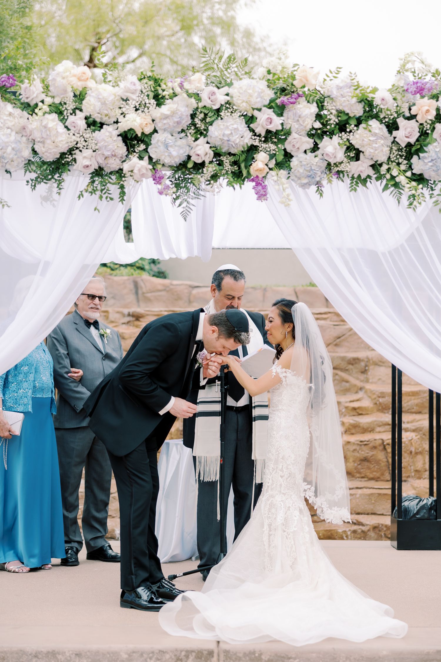 Bride and groom share a kiss under a floral chuppah adorned with white and purple blooms at an outdoor wedding ceremony.