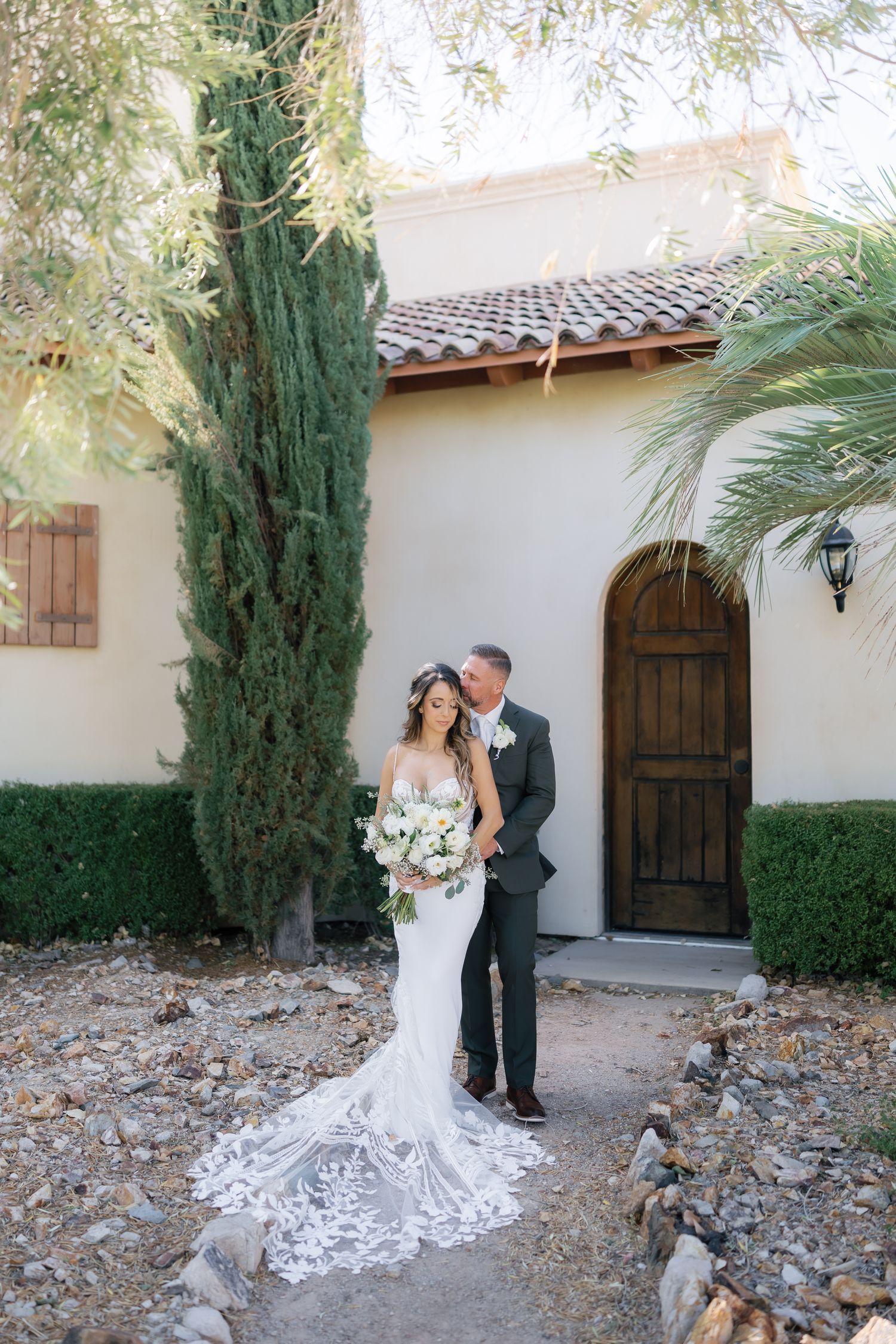 Bride in lace gown holding white bouquet stands with groom in gray suit outside a Spanish-style villa.