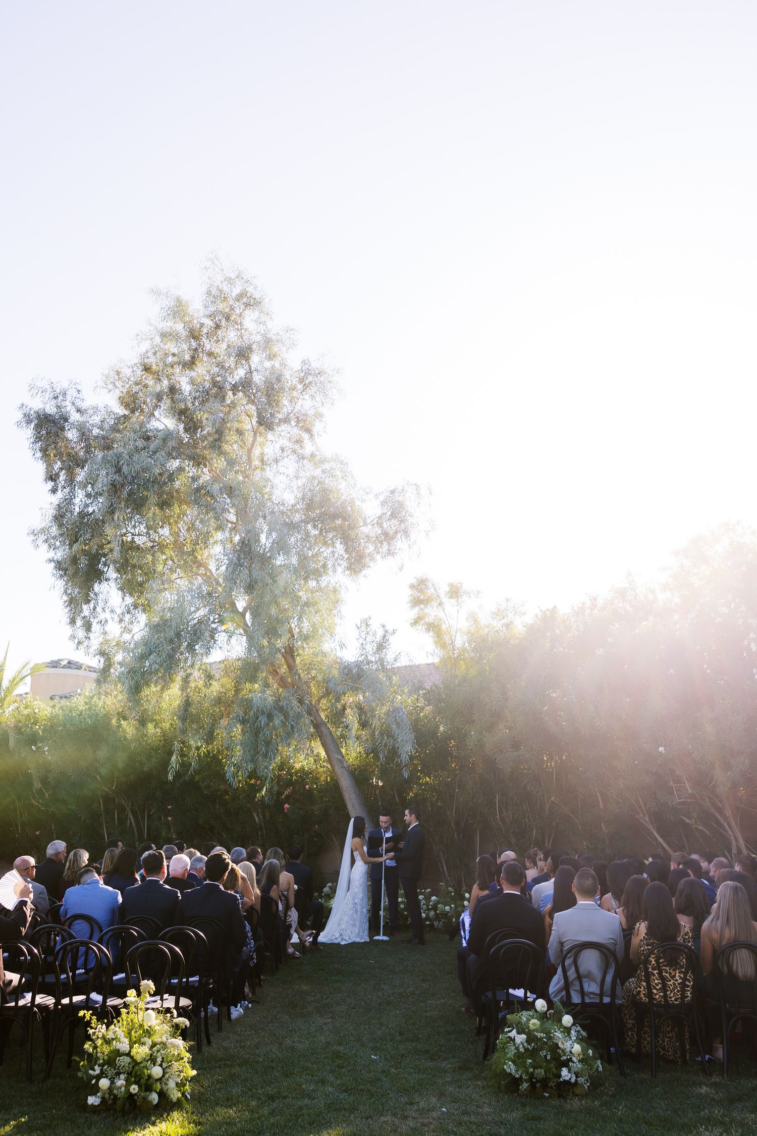 Outdoor garden wedding ceremony with guests seated on chairs and sunlight streaming through tall trees in the background.