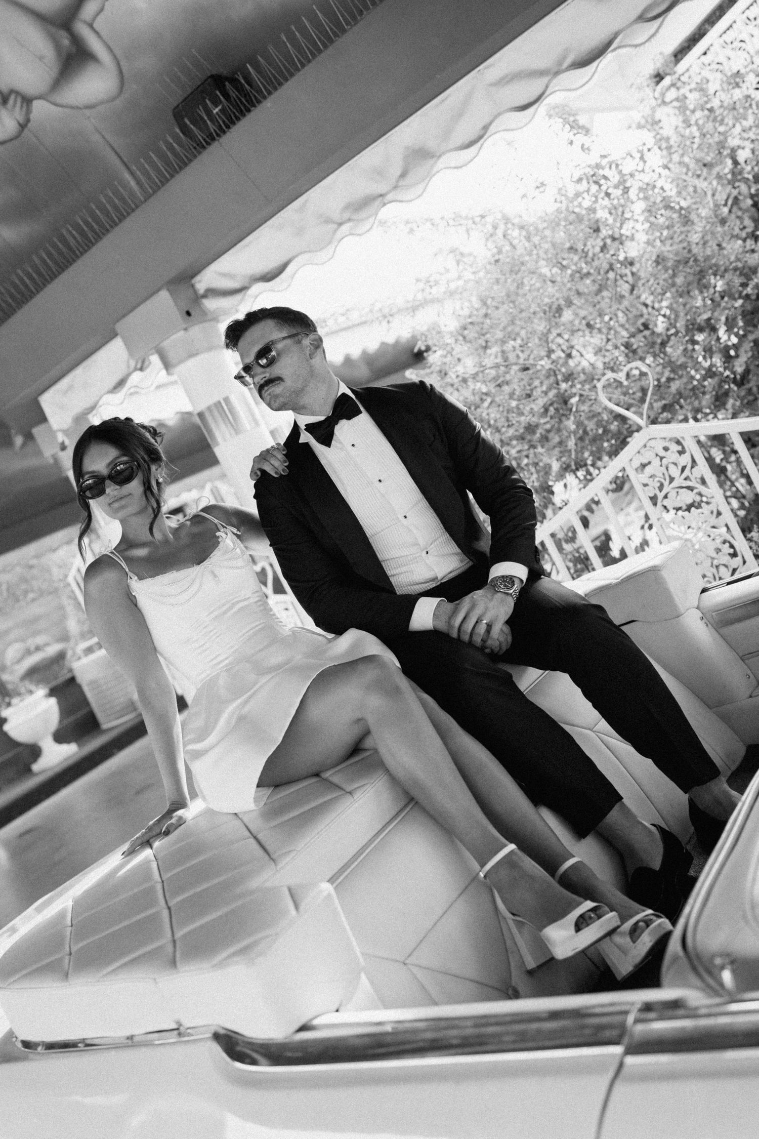 Elegant couple in black and white photo, man in tuxedo and woman in white dress with sunglasses, seated in classic car.