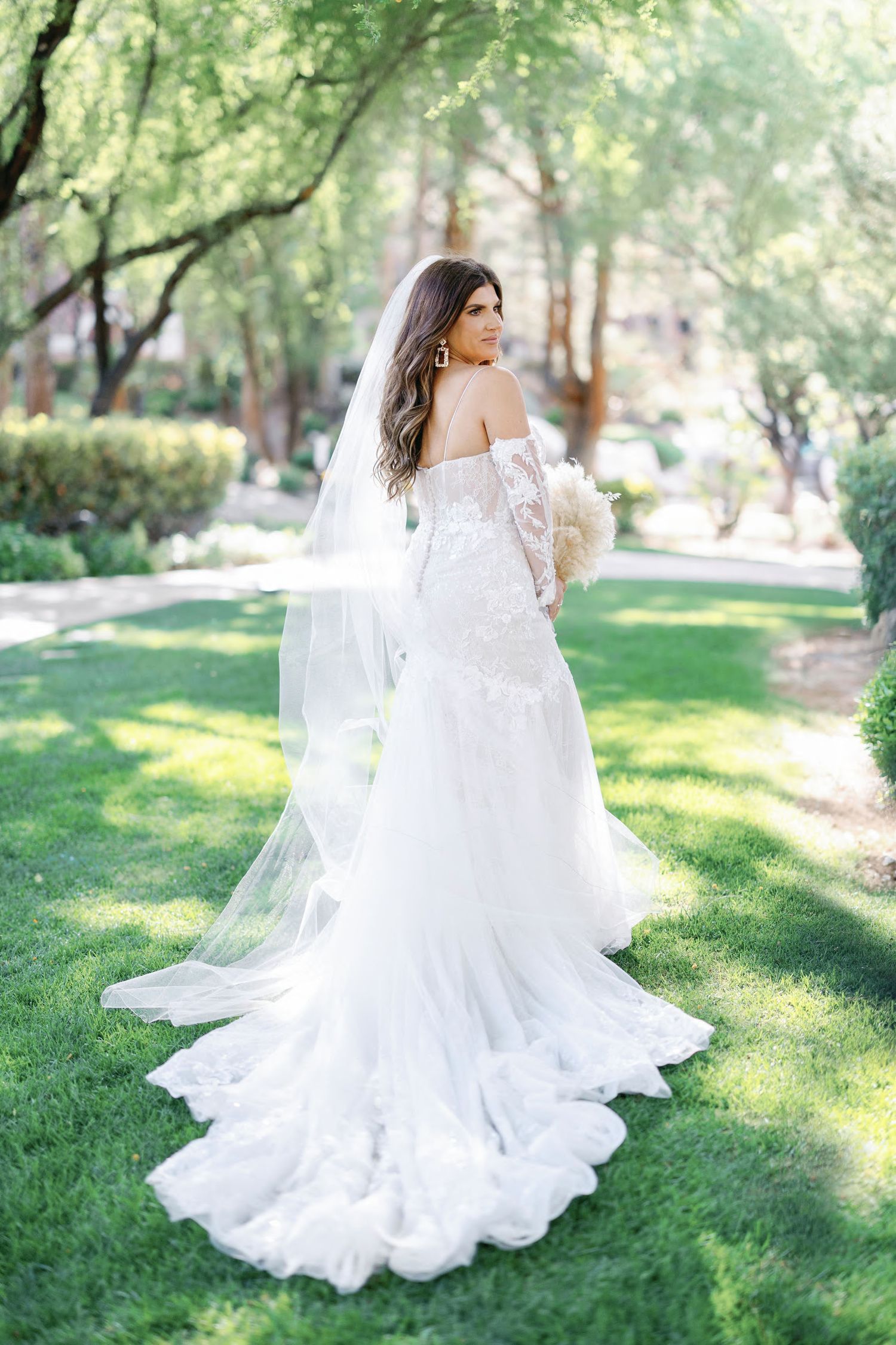 Bride in off-shoulder lace wedding gown with long veil holding bouquet, standing on green lawn surrounded by trees.