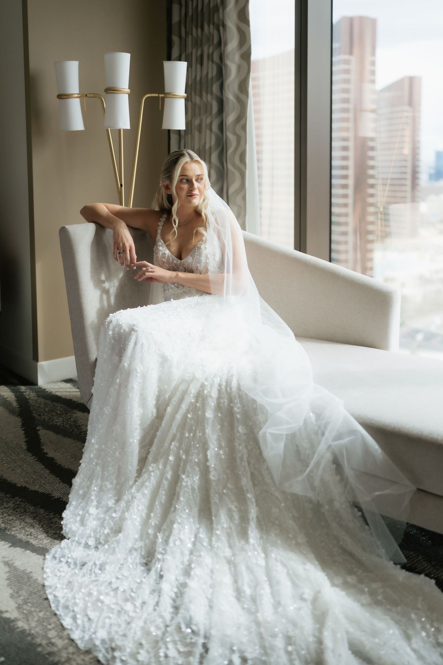 Bride in a sparkling white ball gown and veil sits gracefully in a chair by a large window with city views.