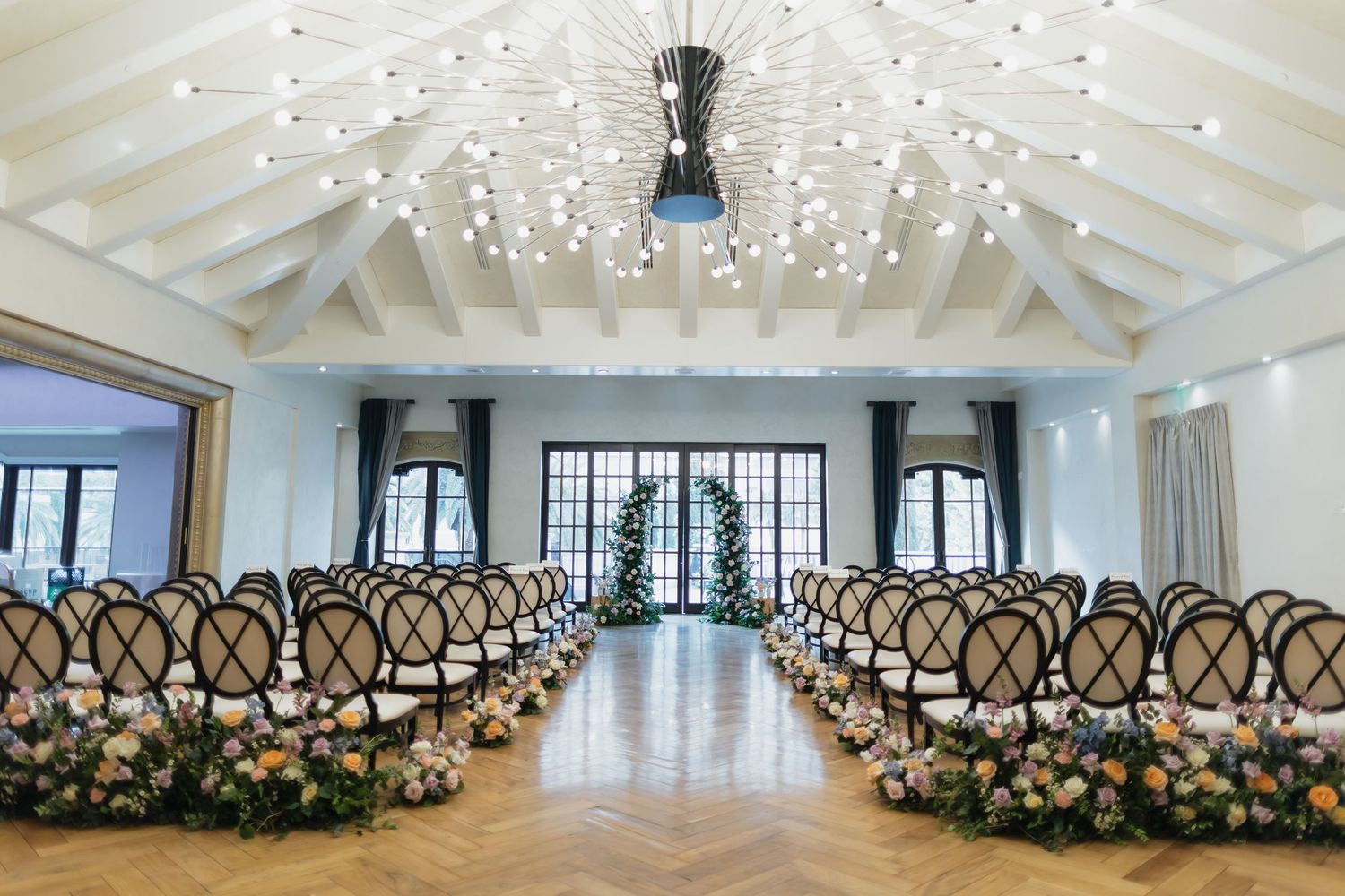Elegant wedding ceremony hall with herringbone floors, floral aisle decor, chiavari chairs, and a chandelier ceiling.