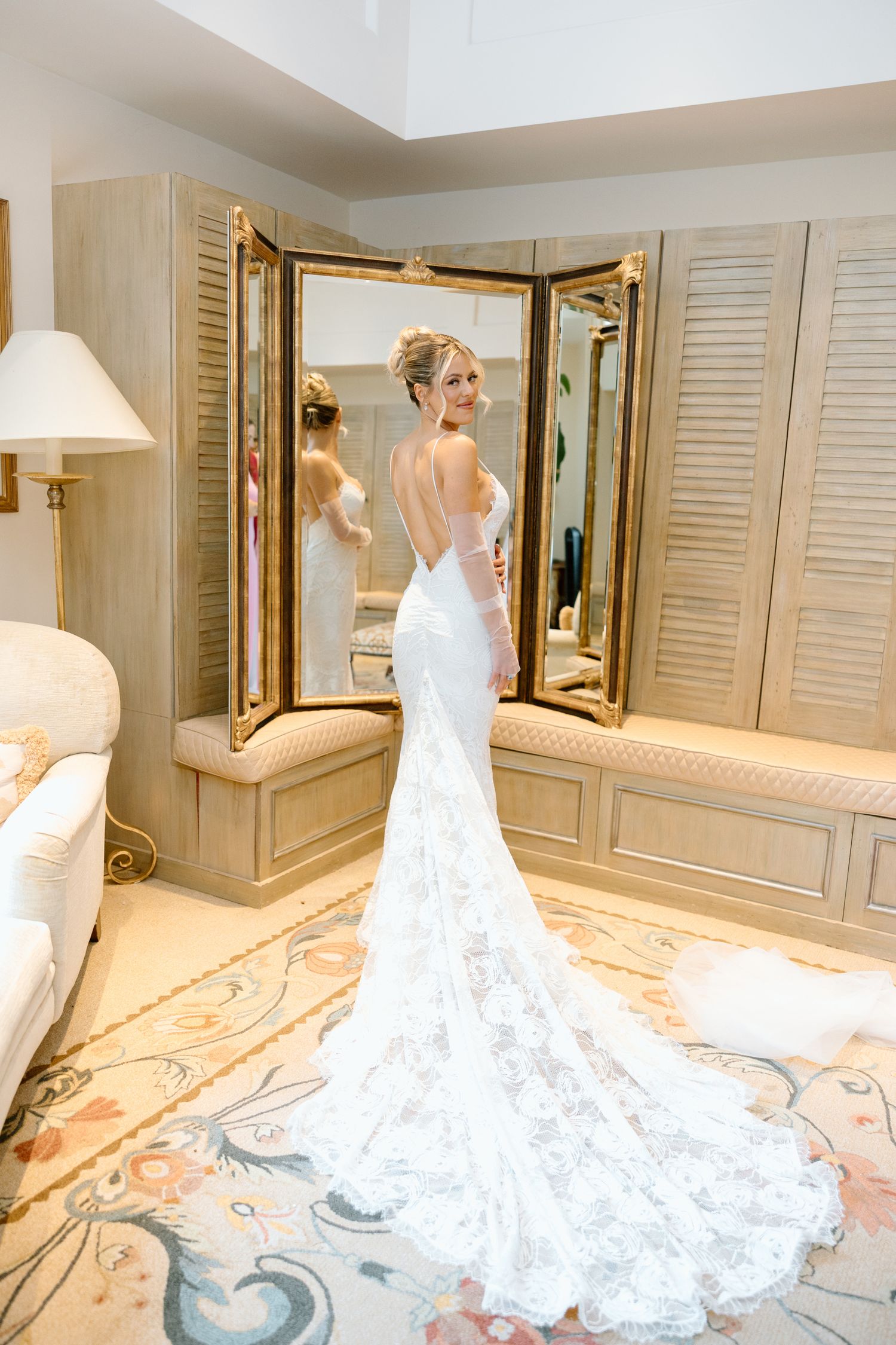 Bride in elegant backless lace wedding gown with train stands before gold-framed trifold mirror in luxury bridal suite.