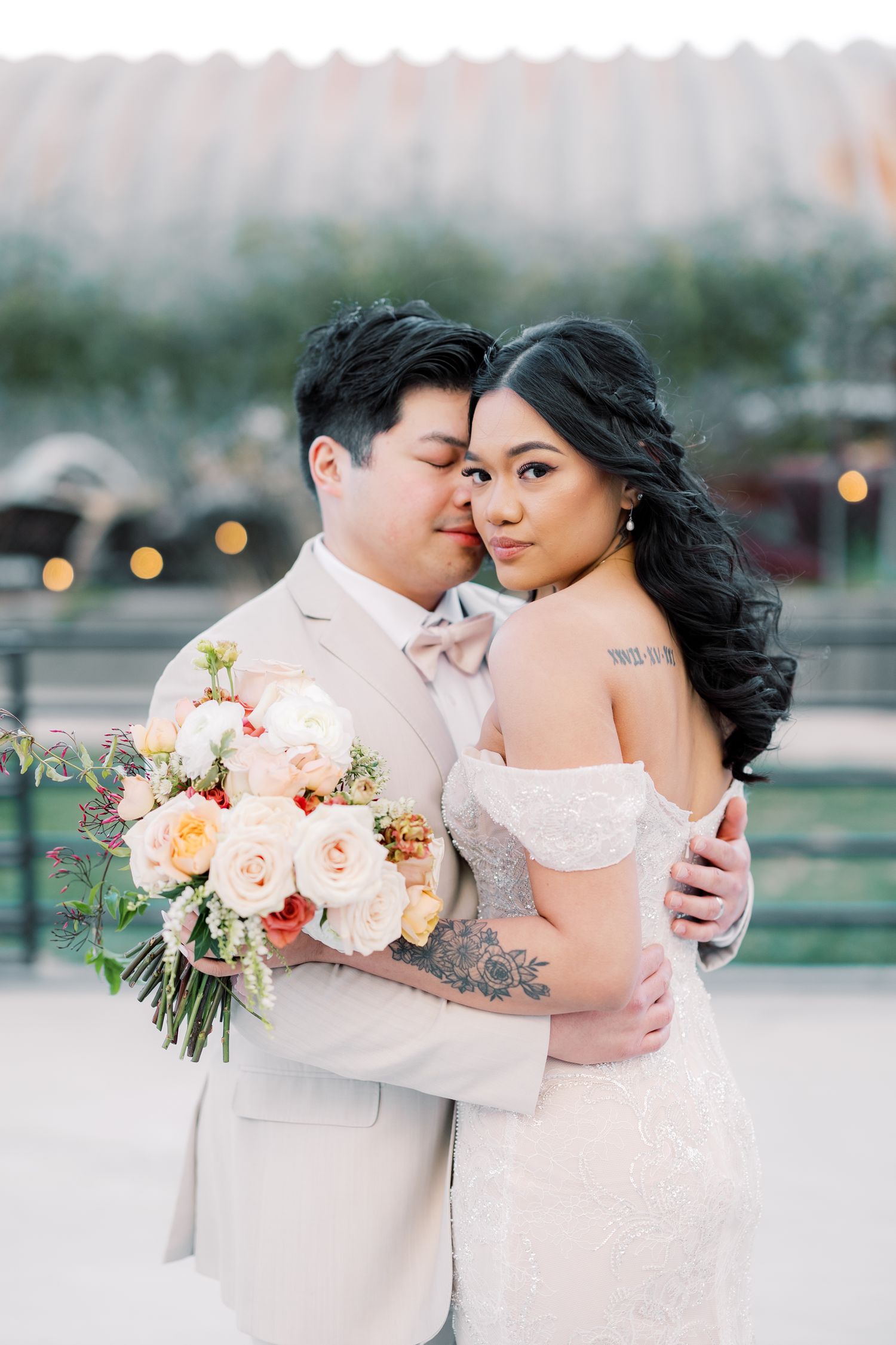 A couple in wedding attire poses with a blush pink and peach floral bouquet against an outdoor evening setting.