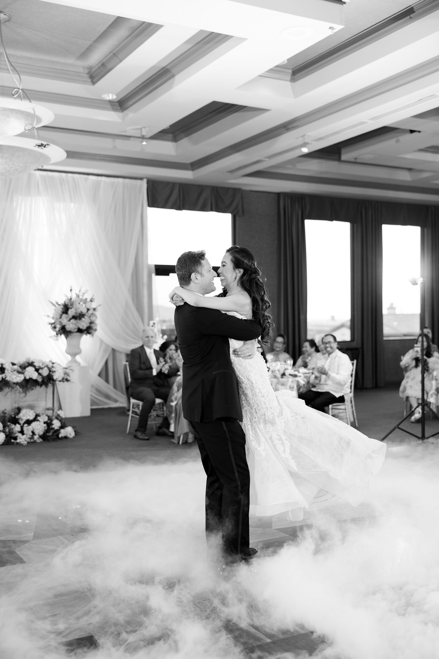 Bride and groom share first dance on fog-covered floor at elegant wedding reception in black and white photo.