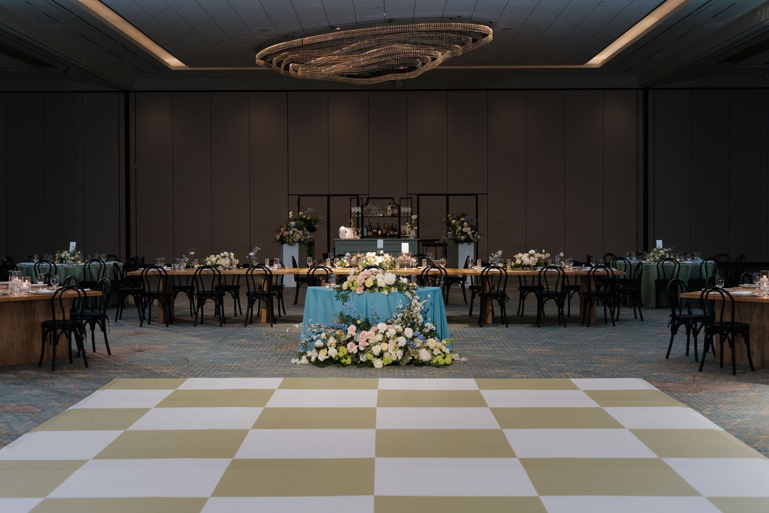 Elegant wedding reception ballroom with checkered dance floor, floral centerpieces, teal linens, and dramatic chandelier lighting.