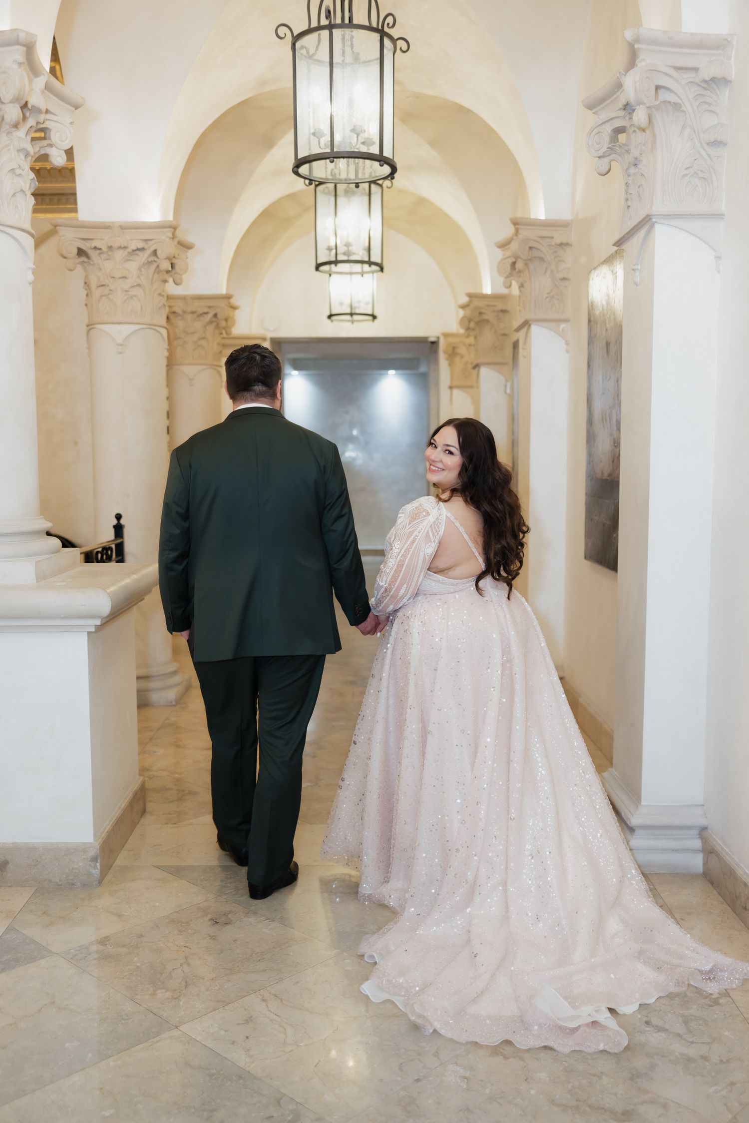 Bride in pink lace gown holds grooms hand walking through marble columned wedding venue hall.