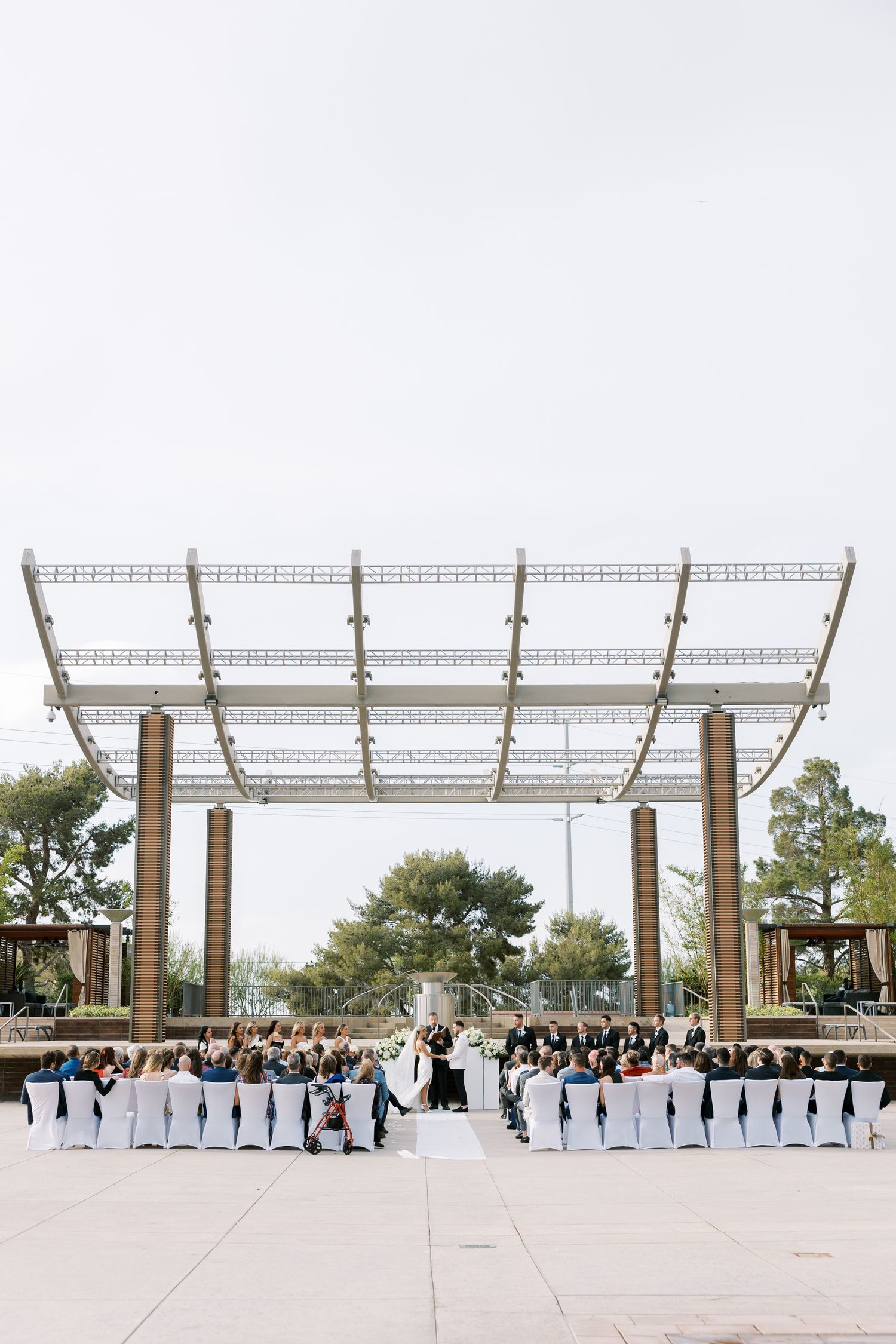 Outdoor wedding ceremony under a modern pergola with guests seated in white chairs on a plaza.