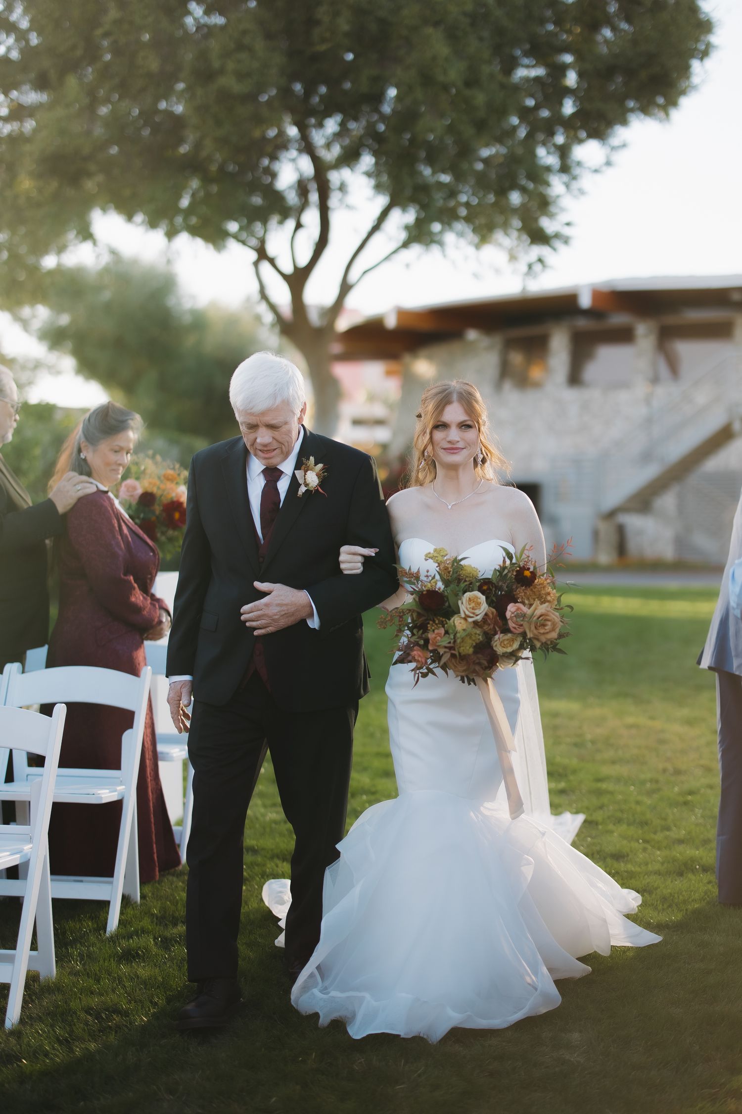 Bride in mermaid gown walks down outdoor aisle with older gentleman escort holding autumn bouquet.