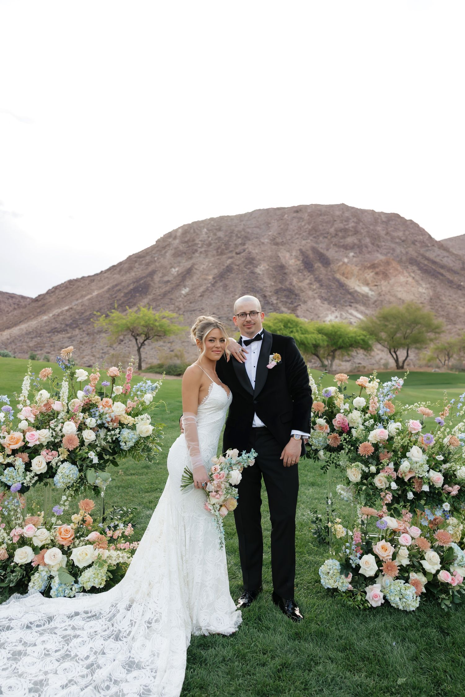 Bride and groom pose before floral arrangements with desert mountain backdrop on lush green lawn.
