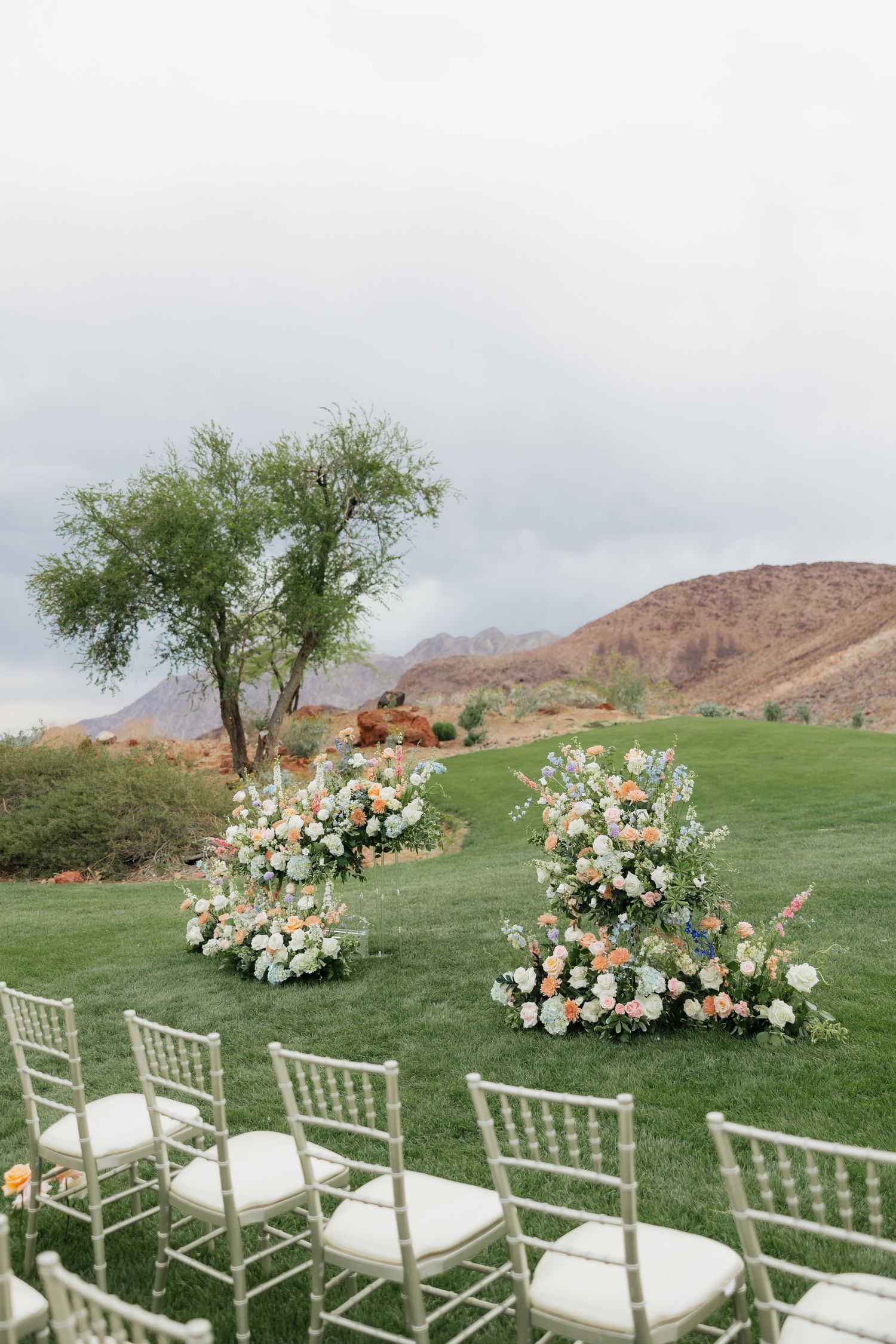 Outdoor wedding ceremony setup with floral arrangements on green lawn, chiavari chairs, and mountain backdrop under cloudy sky.