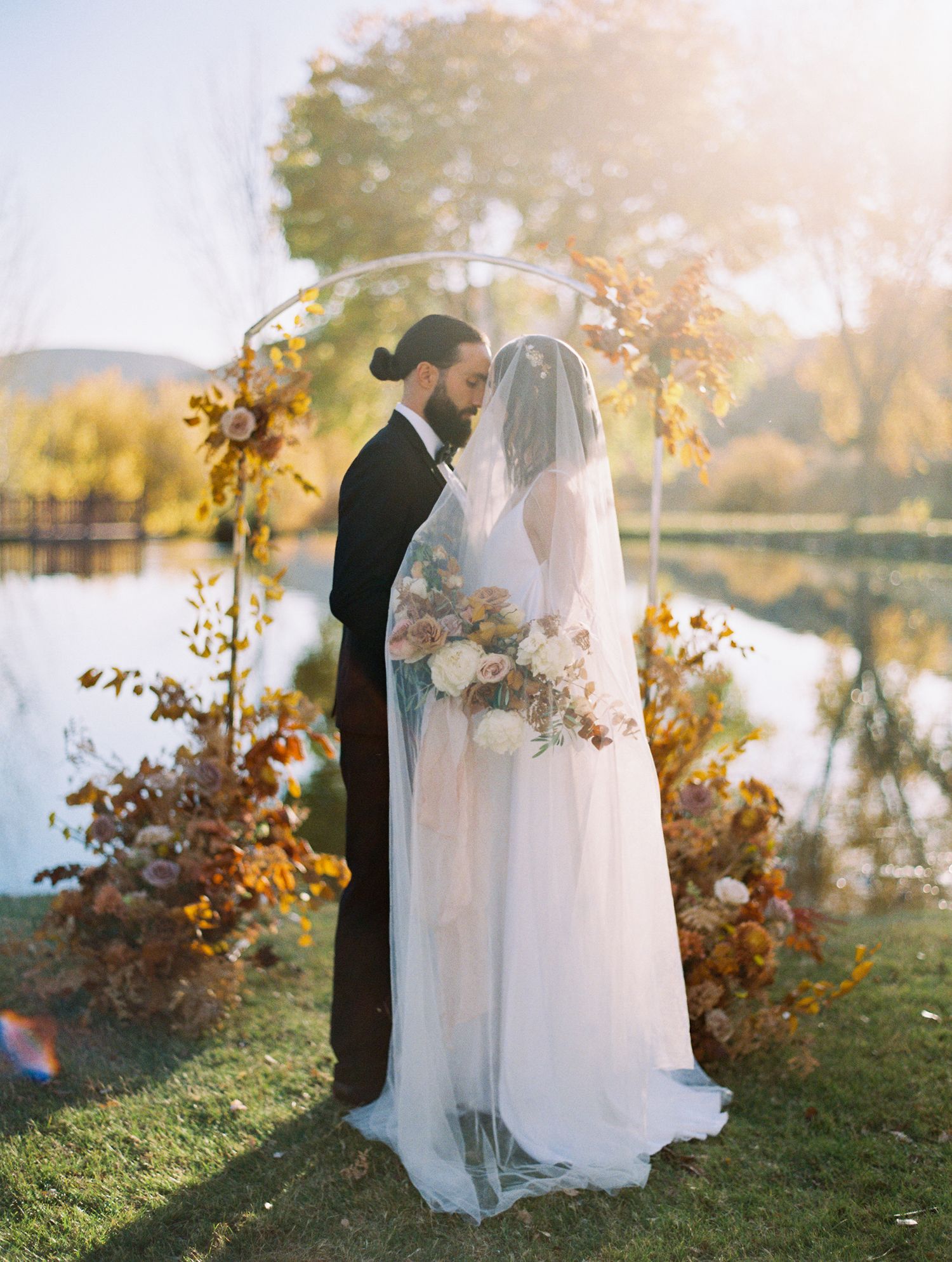 A couple in wedding attire embraces by a lake during golden hour with autumn foliage in the background.