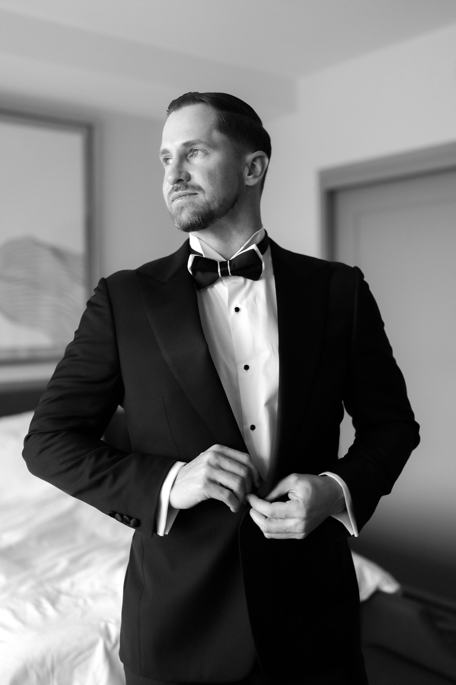 Black and white photo of a groom in a tuxedo and bow tie buttoning his jacket in a hotel room.