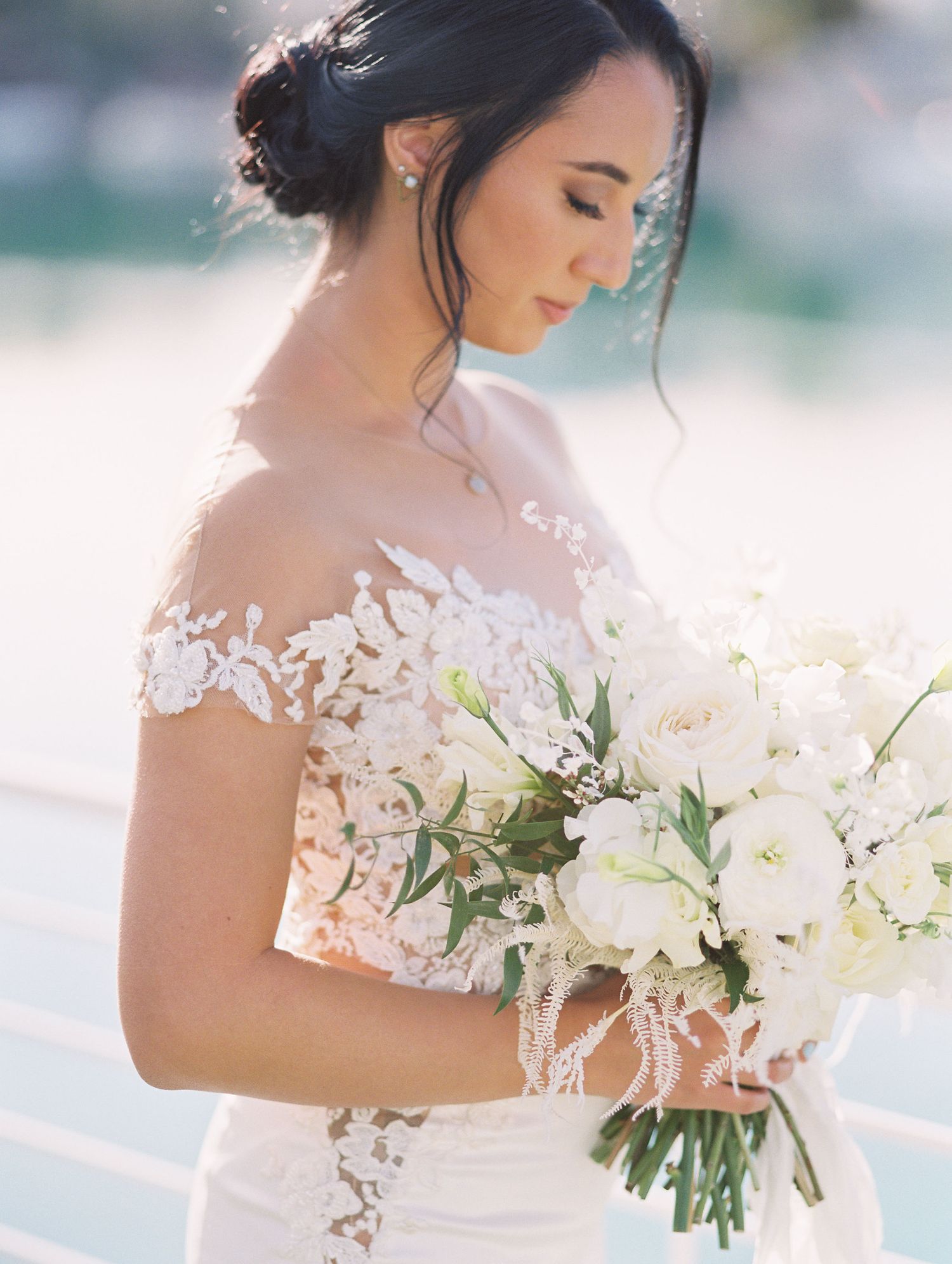 Bride in lace off-shoulder wedding gown holds white floral bouquet with roses and greenery in soft natural light.