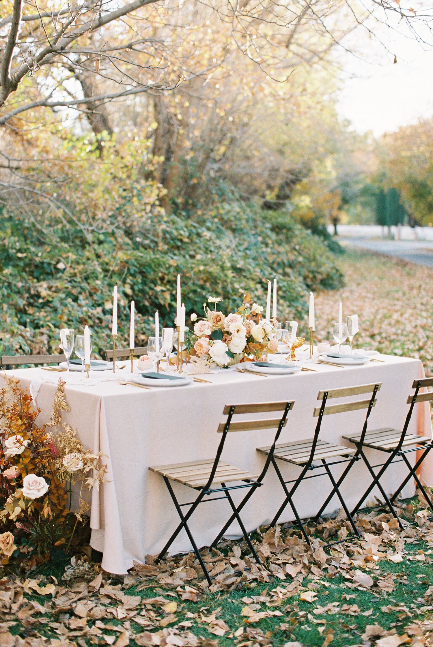 Elegant outdoor fall wedding table setting with white linens, candles, floral centerpiece, and folding chairs among autumn leaves.