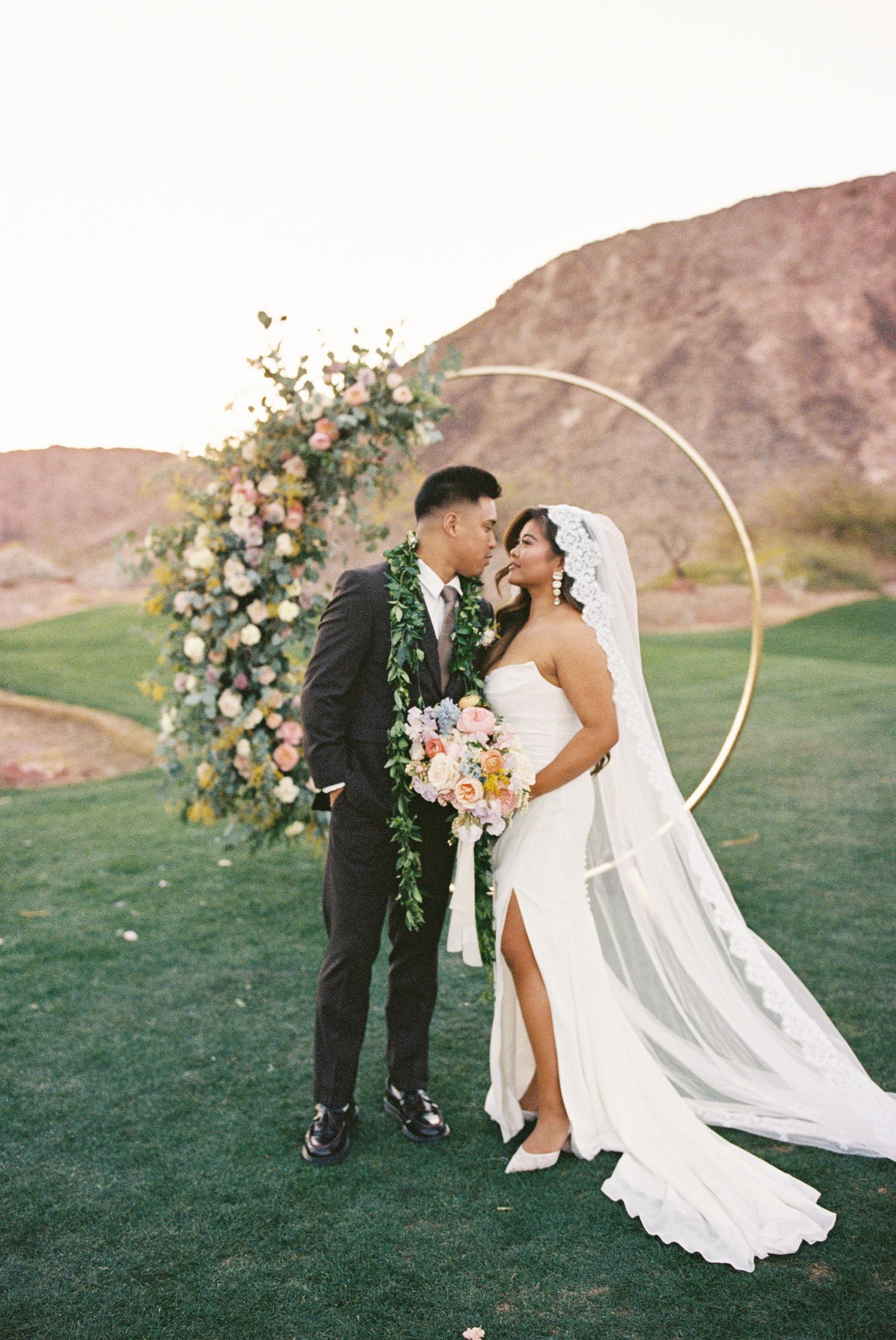 Bride and groom share a romantic moment in front of a circular floral arch on a golf course with red rock mountains behind.