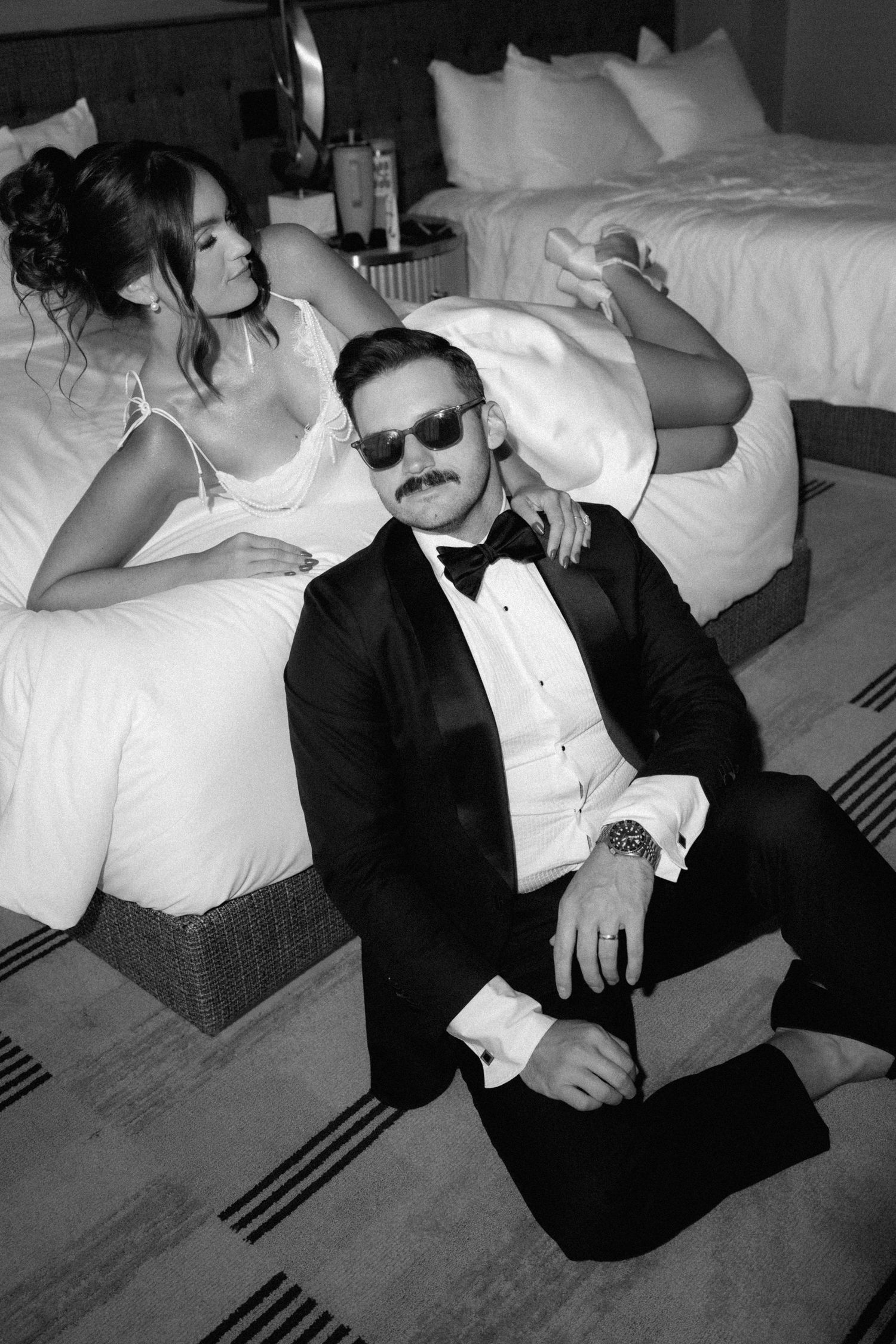 Black and white photo of a groom in tuxedo sitting on floor while bride in white dress poses on hotel bed.