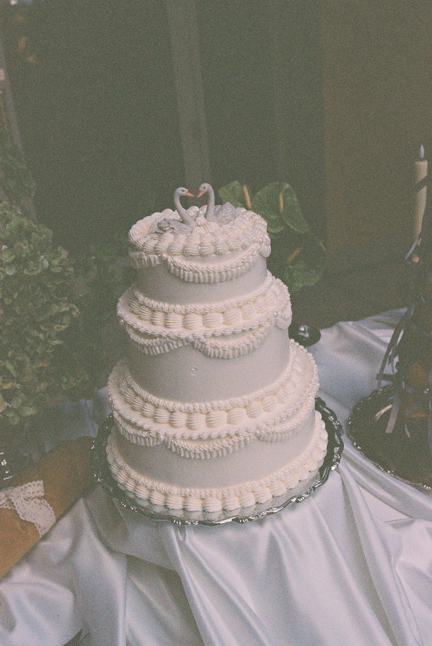 Three-tier white wedding cake with pearl borders and swan topper on satin draped table.