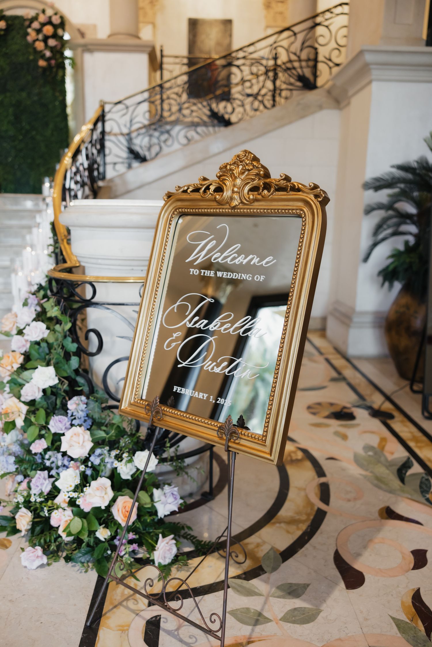 Gold ornate mirror wedding welcome sign on easel with floral arrangement at elegant venue staircase.