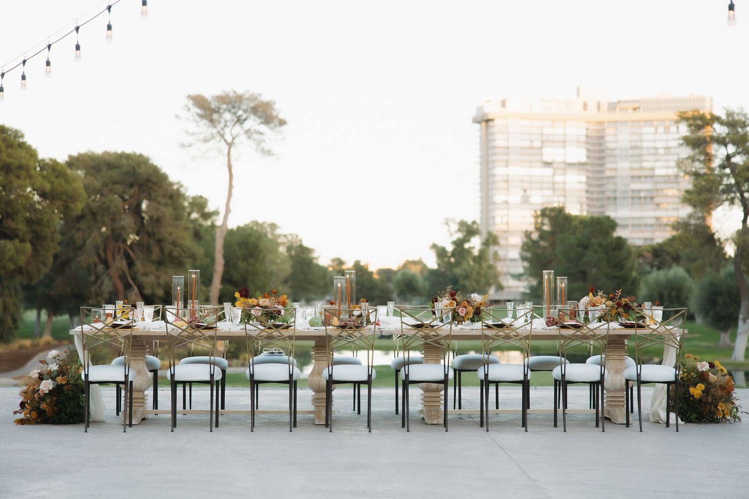 Elegant outdoor wedding reception with long dining table, floral arrangements, and city backdrop at dusk.