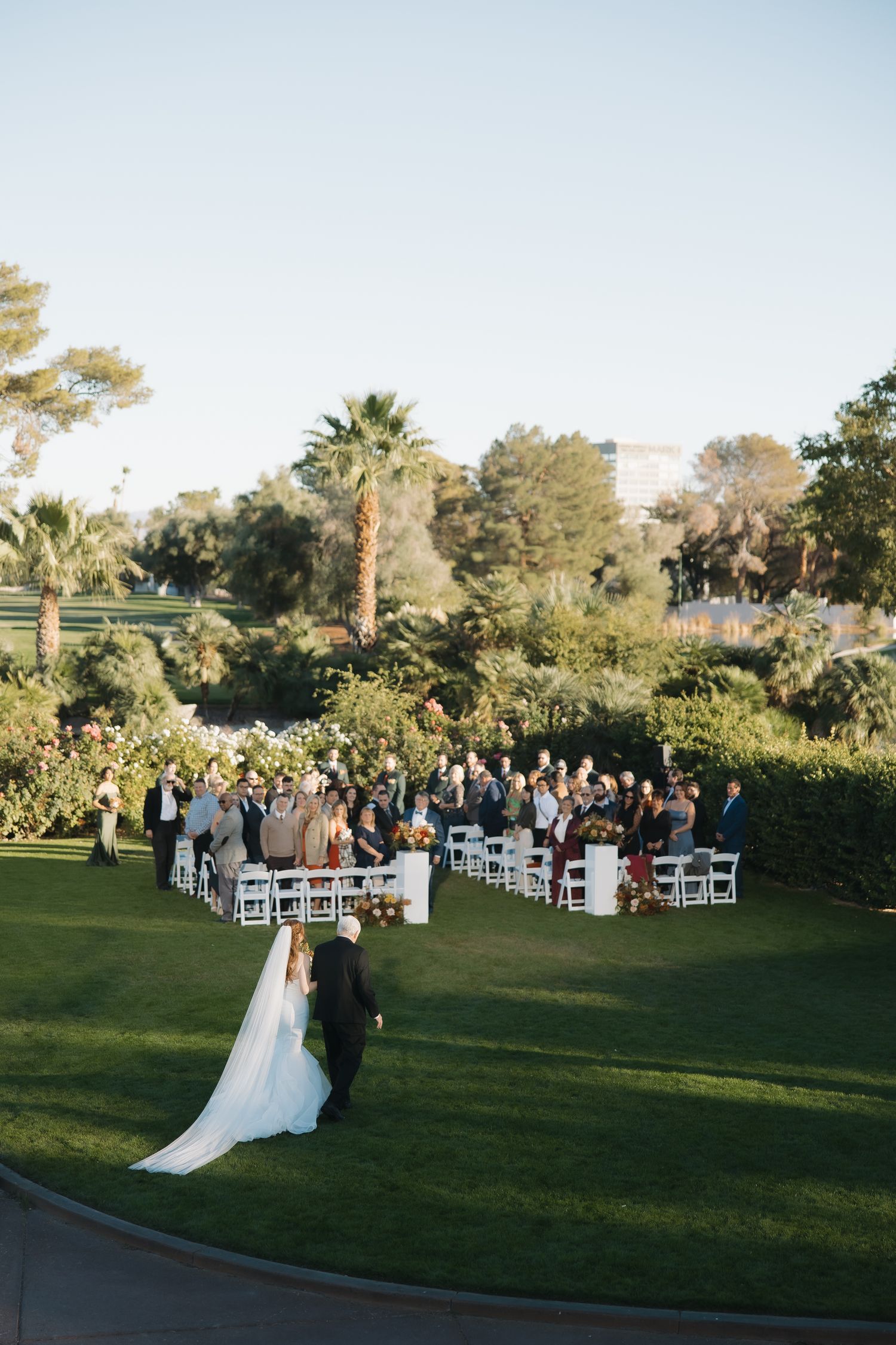 Bride and groom walk toward wedding guests seated on white chairs on a lush green lawn.