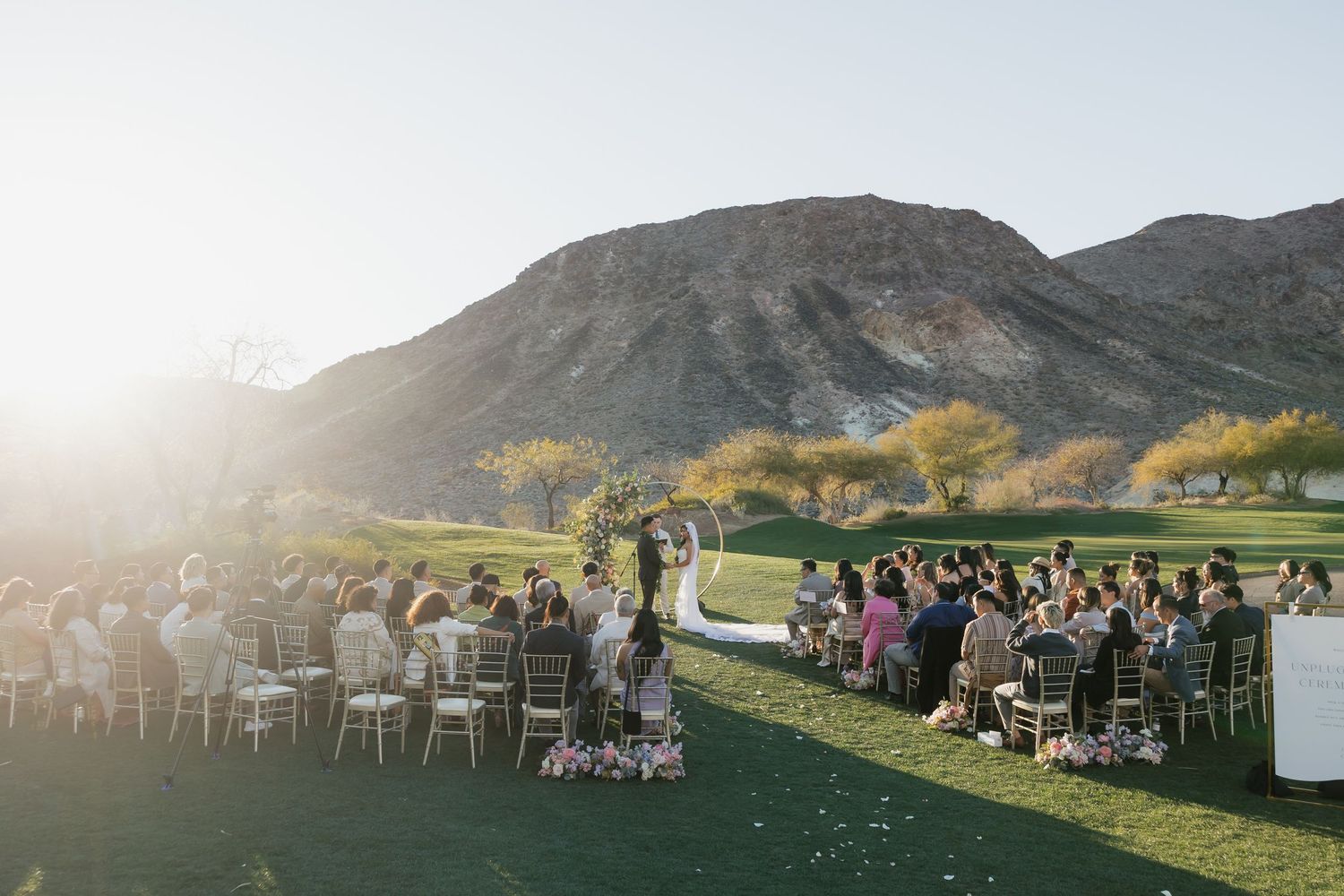 Outdoor wedding ceremony on lush green lawn with mountain backdrop at sunset in Palm Springs desert venue.