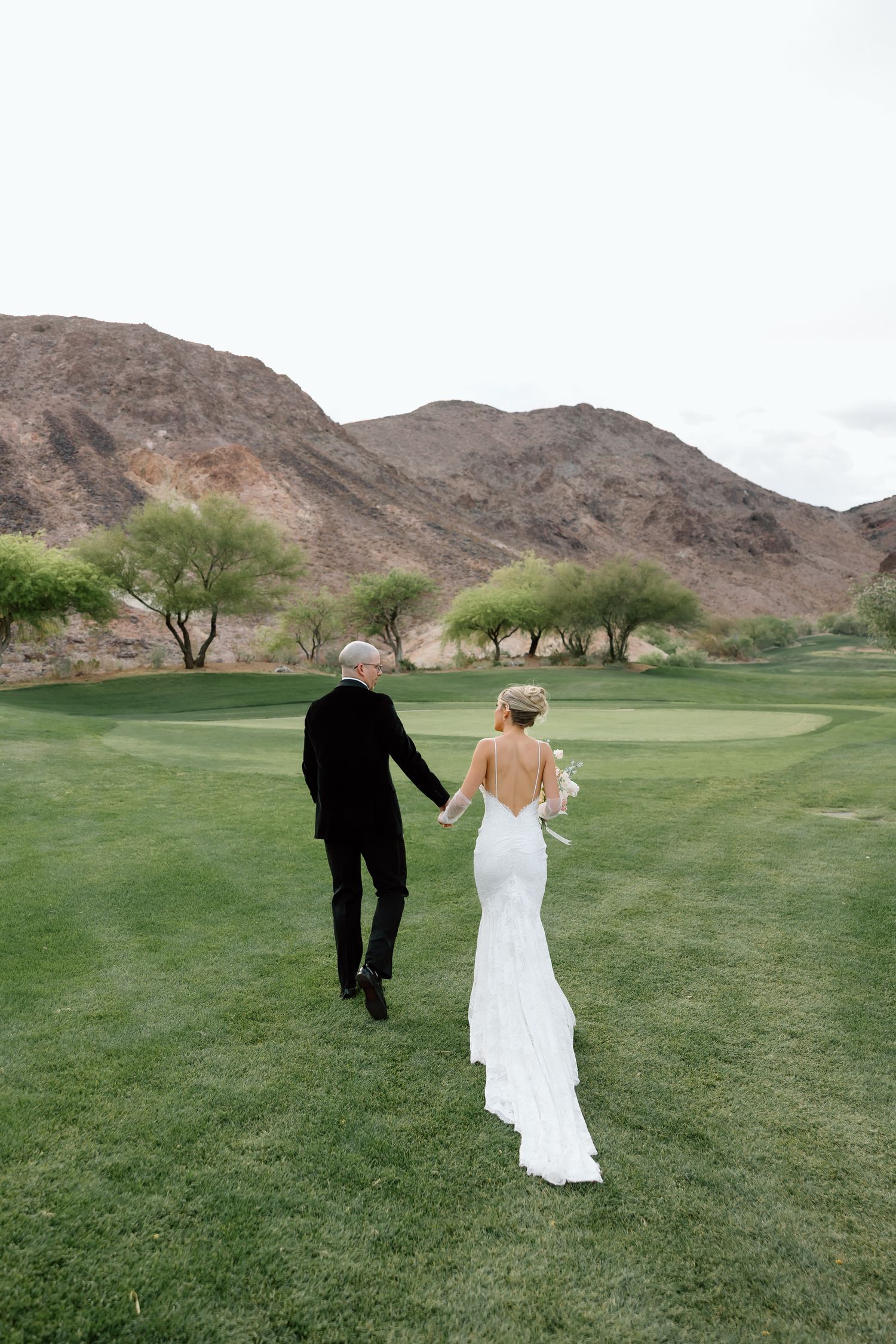 Bride and groom walk hand-in-hand across lush golf course fairway with desert mountains backdrop.