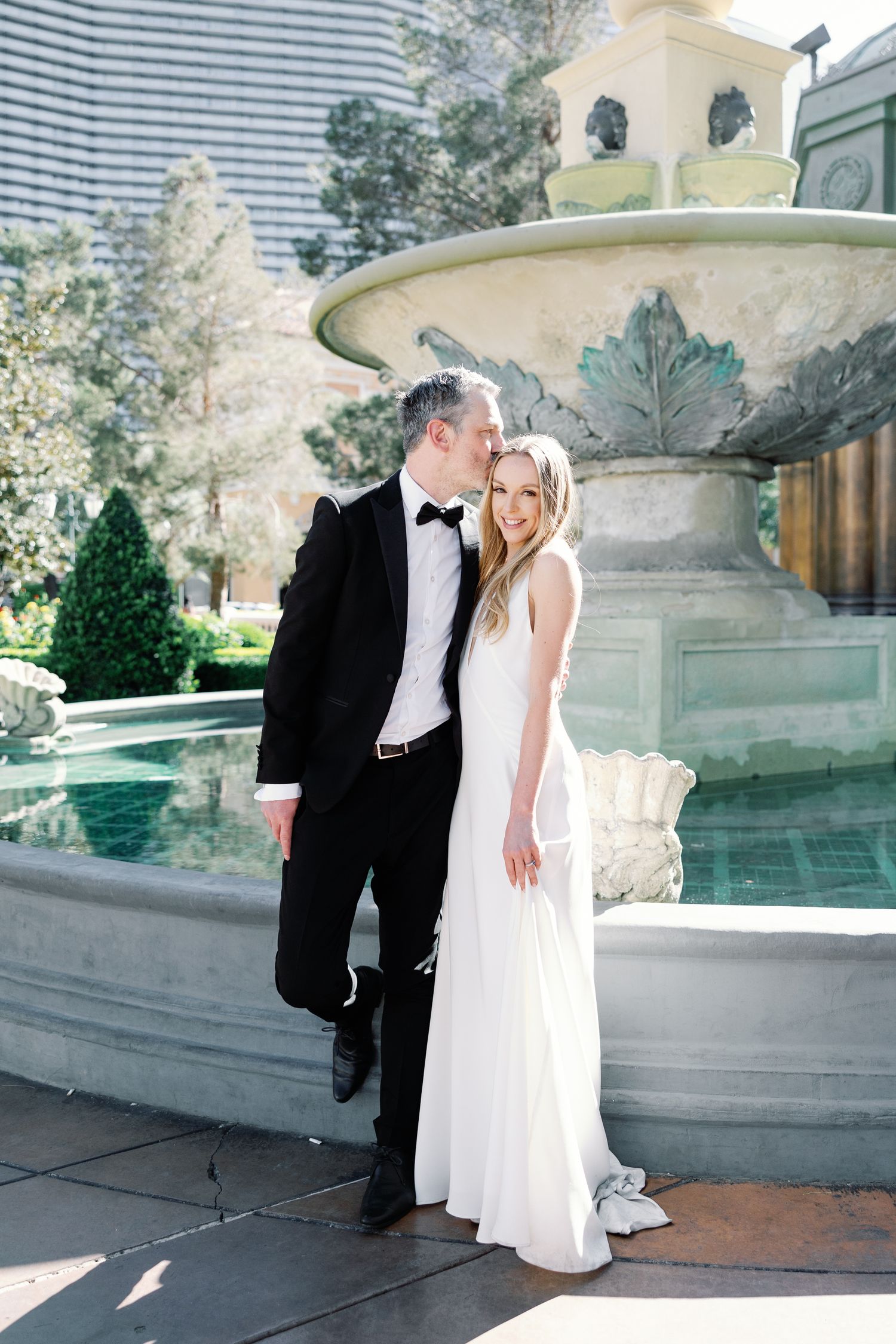 A bride and groom pose together in front of an elegant fountain, she in a white gown, he in a classic black tuxedo.