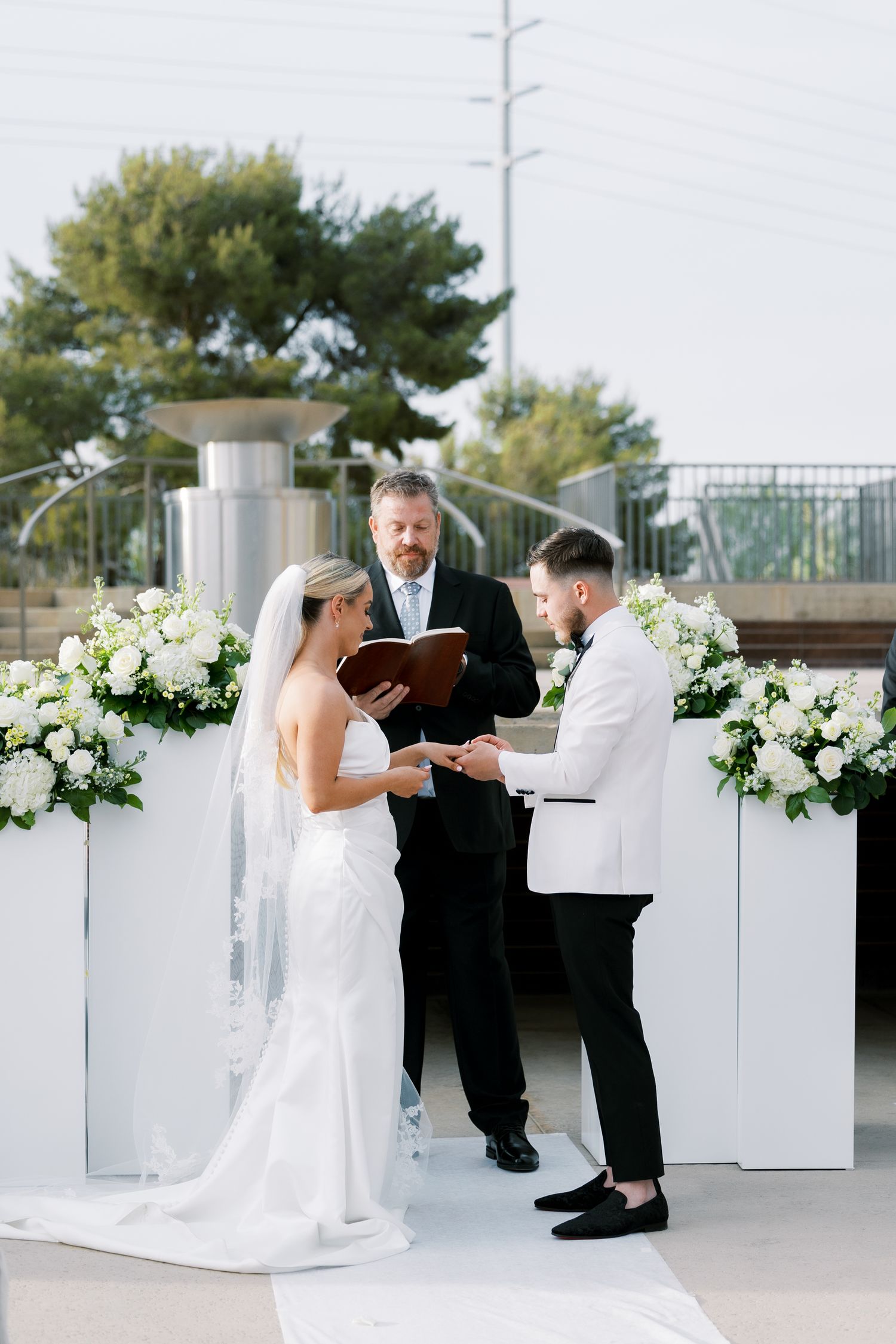 Bride and groom exchange rings during outdoor wedding ceremony, officiated by minister with white floral arrangements.
