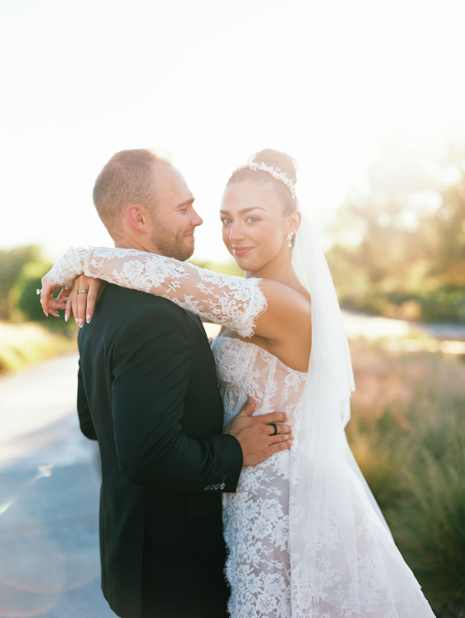 Bride in a lace wedding gown, veil and beaded headpiece embraces her groom in a black suit outdoors in warm golden sunlight.
