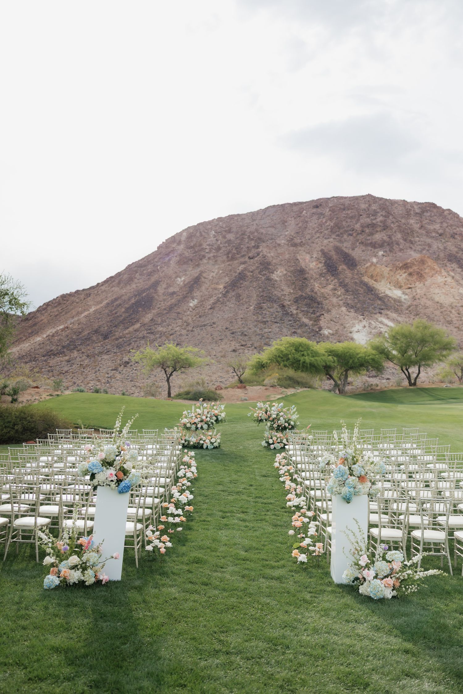 Elegant outdoor wedding ceremony setup on green lawn with floral-lined aisle, white chairs, and scenic desert mountain backdrop.