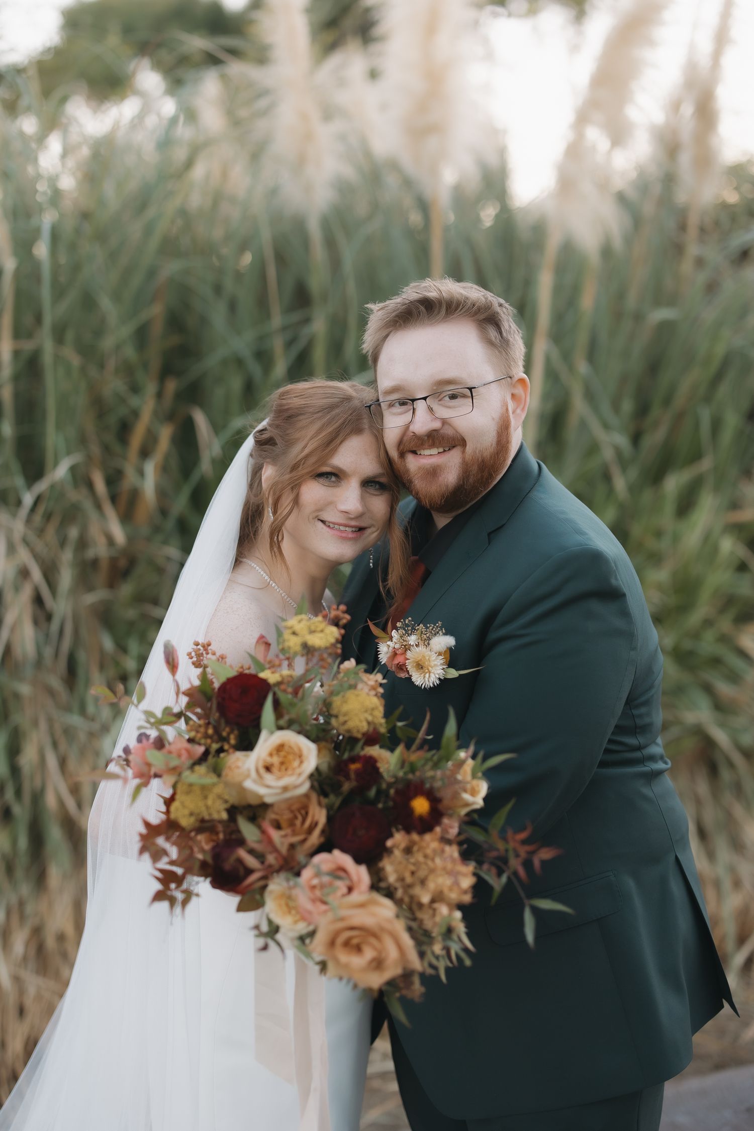 Smiling bride and groom pose together holding lush autumn floral bouquet in front of tall grass.