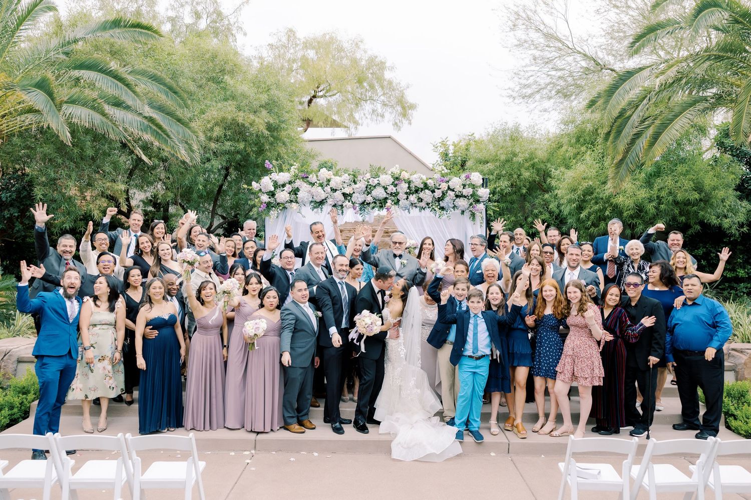 Wedding party and guests pose together outdoors at a garden ceremony with floral arch backdrop.
