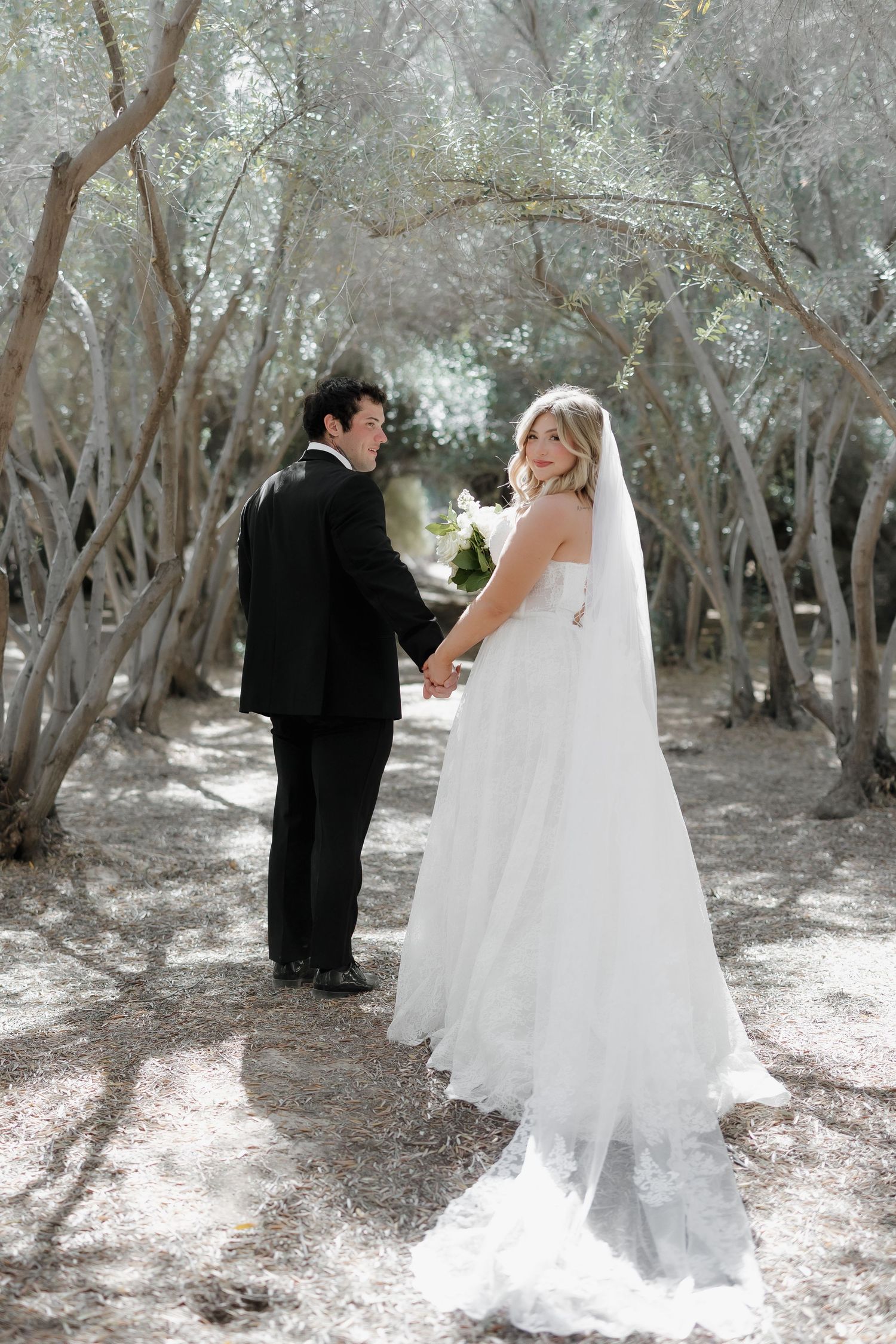 Bride and groom hold hands on a tree-lined path, bride in white gown with veil looks back at camera.