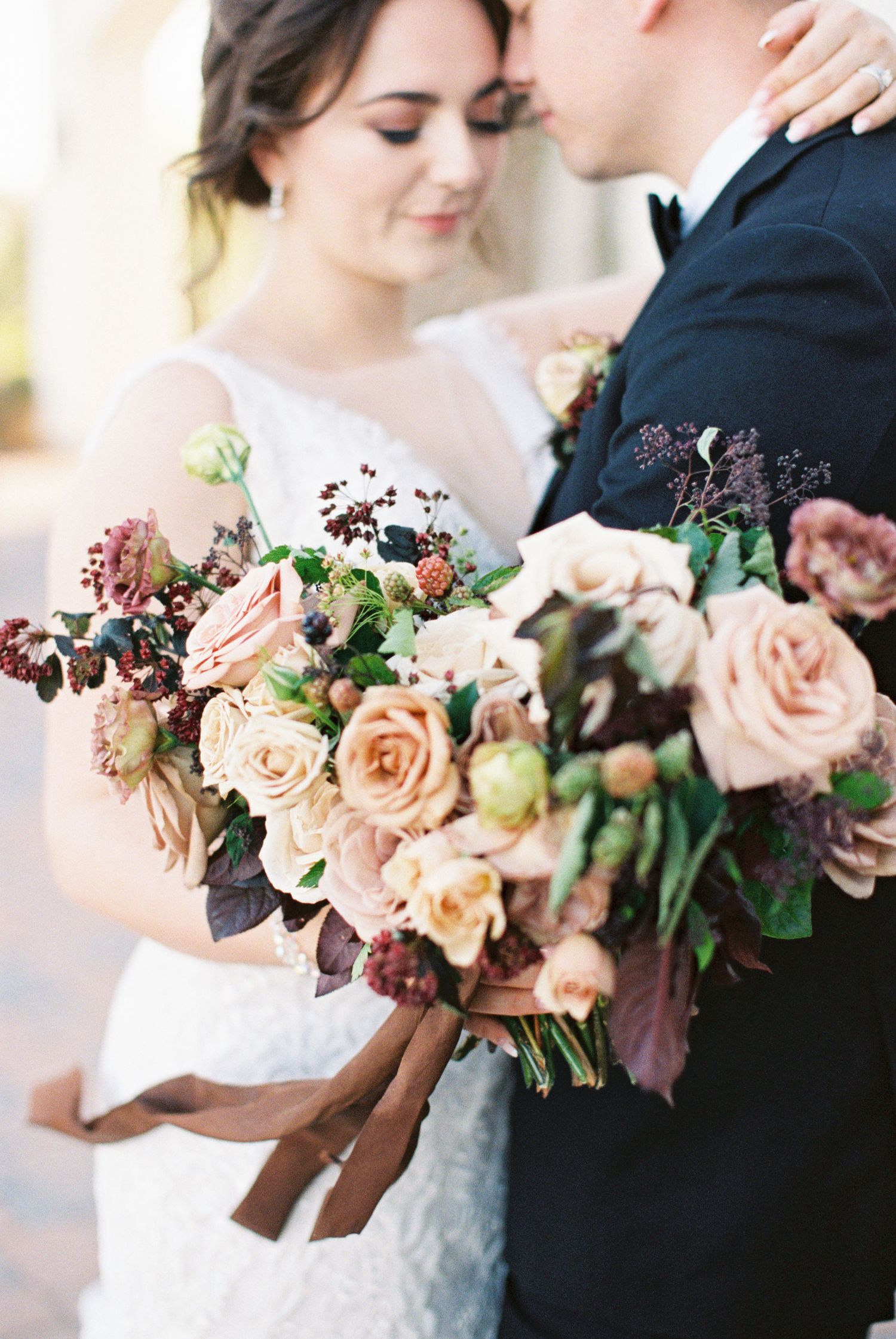 Bride holding lush blush and burgundy rose bouquet with ribbon, standing close to groom in navy suit.