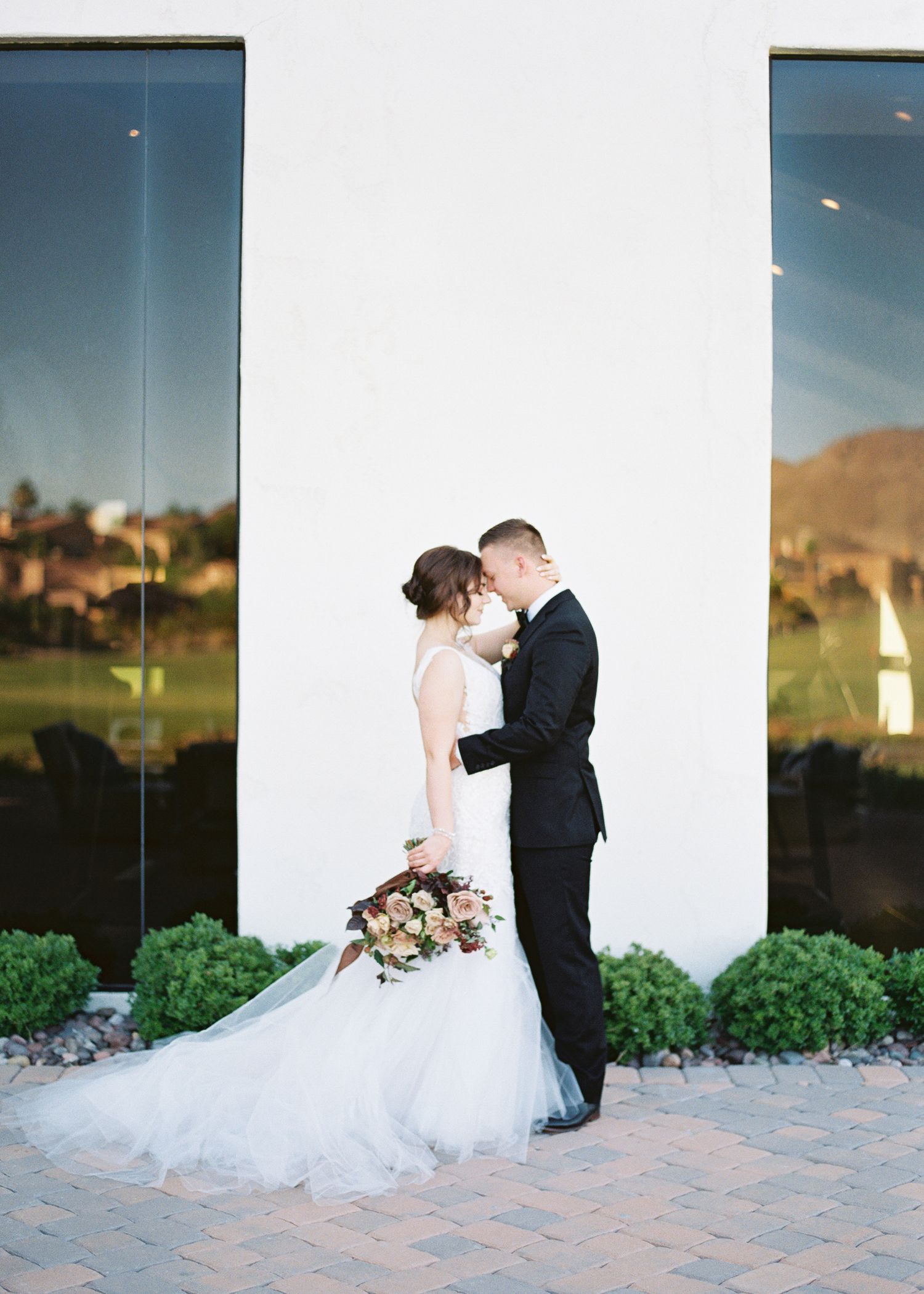 Bride in white tulle gown and groom in black tuxedo share a romantic kiss outdoors against a white wall.