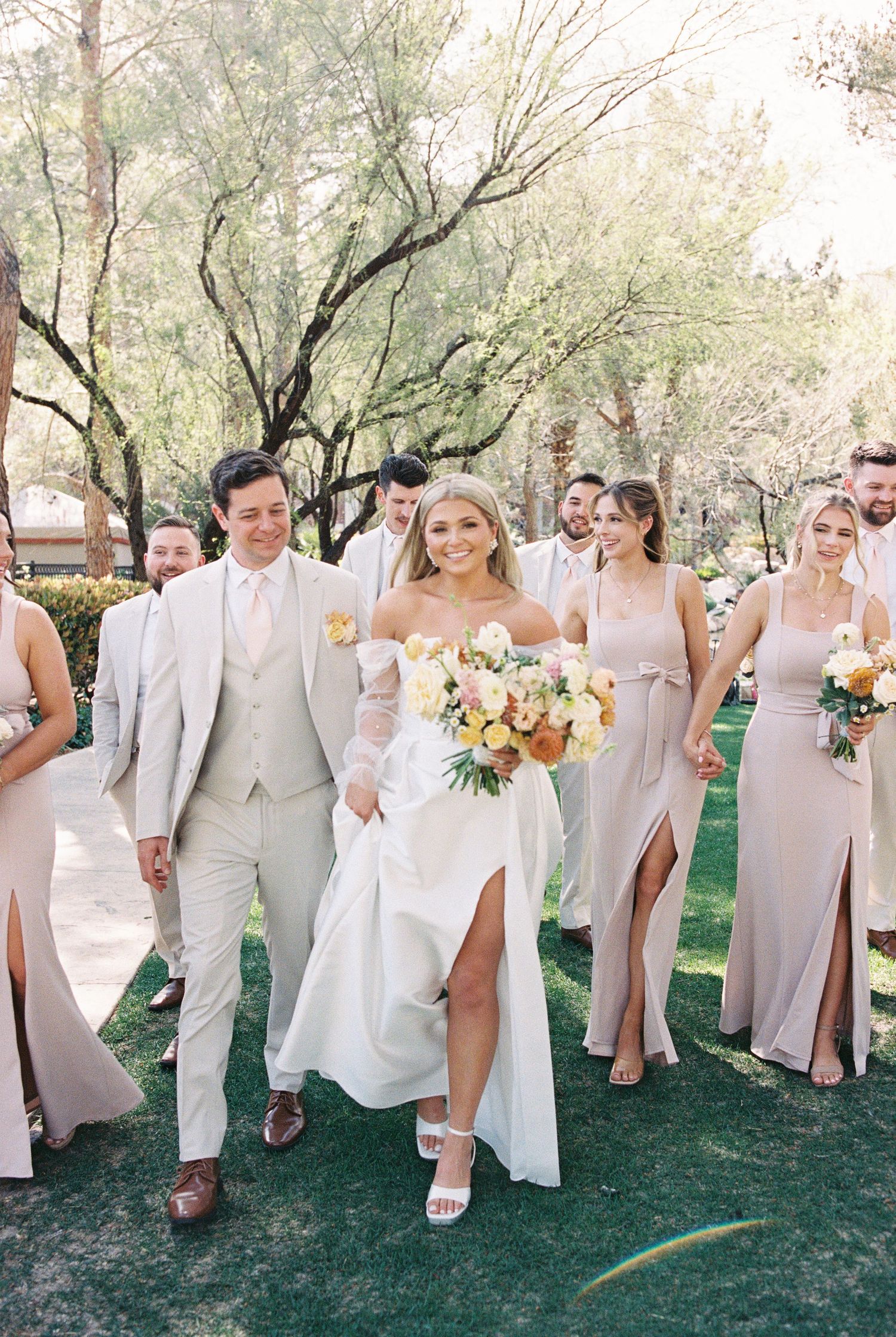 Bride and groom walk with wedding party in neutral tones outdoors, bride holding colorful bouquet.
