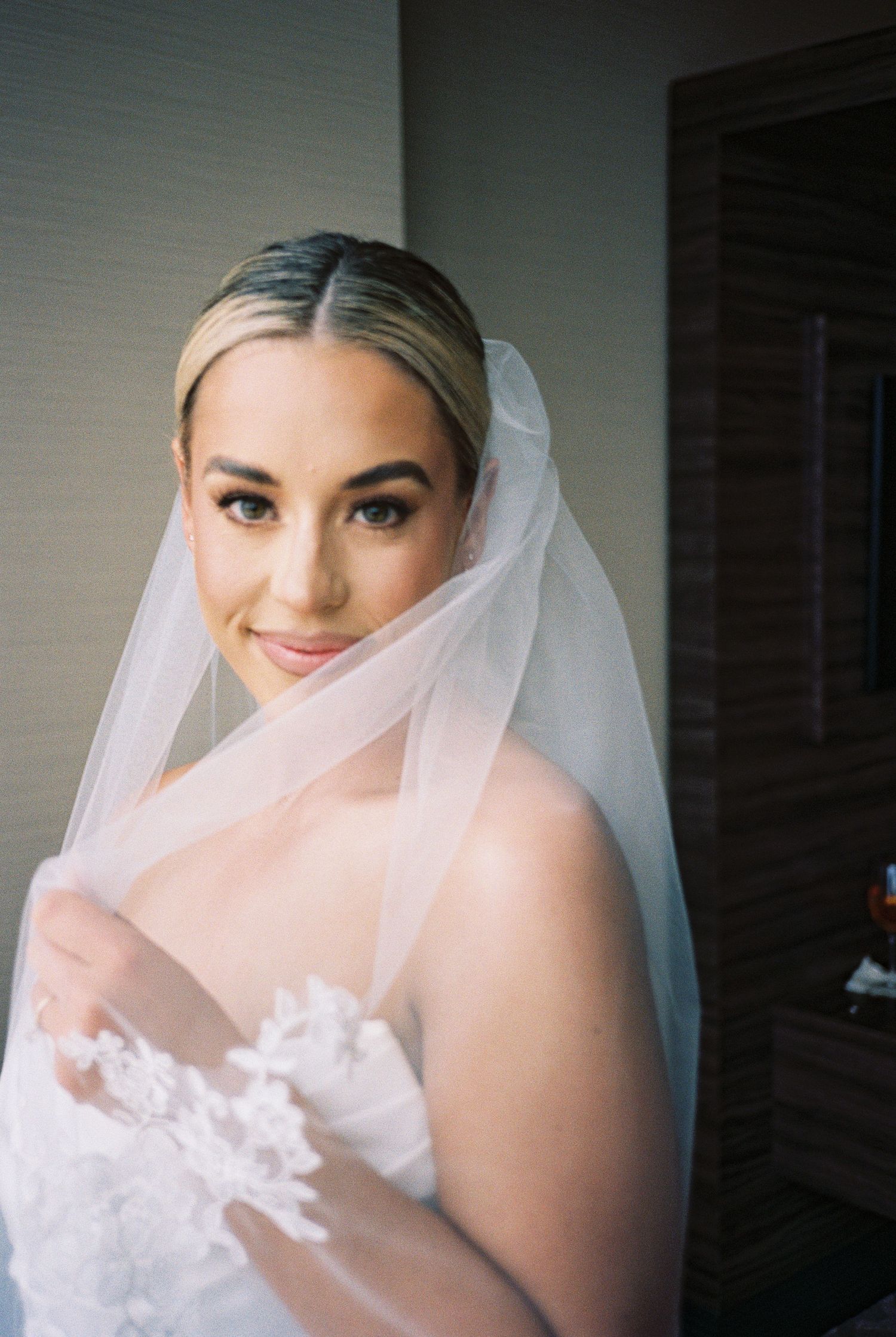 Elegant bride in a lace wedding dress and sheer veil, smiling softly in a beautifully lit indoor setting.